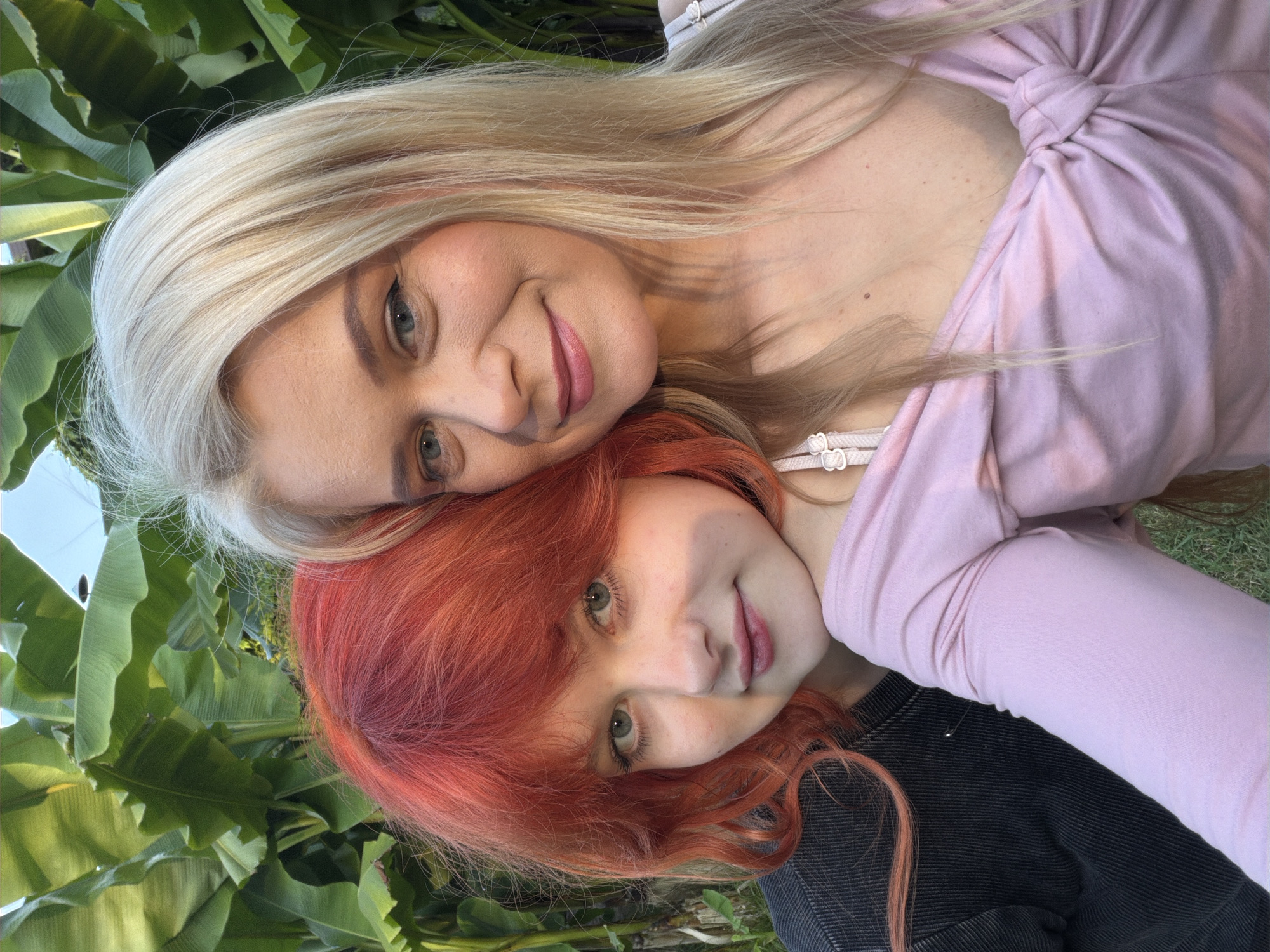 Two individuals are captured in a close-up selfie, posed in front of a backdrop of lush green banana leaves. The person on the left has vibrant red hair and is wearing a dark, ribbed long-sleeved shirt with a small safety pin visible. They are leaning their head against the other person, with their eyes looking directly at the camera. The person on the right has blonde hair and is wearing a light pink, long-sleeved shirt with a knotted detail at the front. They are smiling at the camera, with their gaze slightly off to the side. The lighting suggests it is daytime, with shadows cast on the right person's chest. The overall setting appears to be outdoors, possibly in a garden or park, with no specific identifiable landmarks of Sochi, Russia visible in the frame.