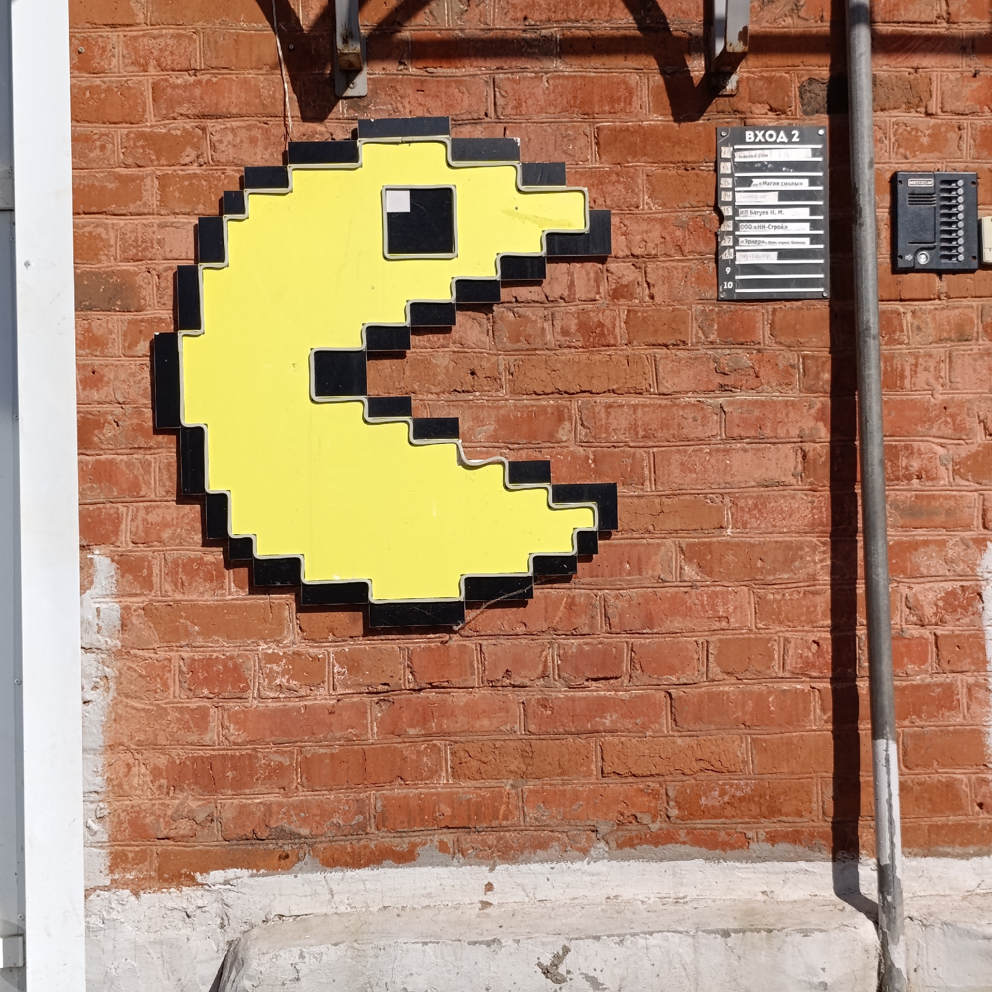 The image shows a brick wall in Chaikovsky, Russia, on which a yellow Pac-Man icon is mounted. Adjacent to the Pac-Man icon are a directory board labeled "ВХОД 2" (Entrance 2) with several business names listed and a gray intercom panel. A vertical gray pipe runs along the wall to the right of the intercom. The wall has a foundation of whitewashed concrete.