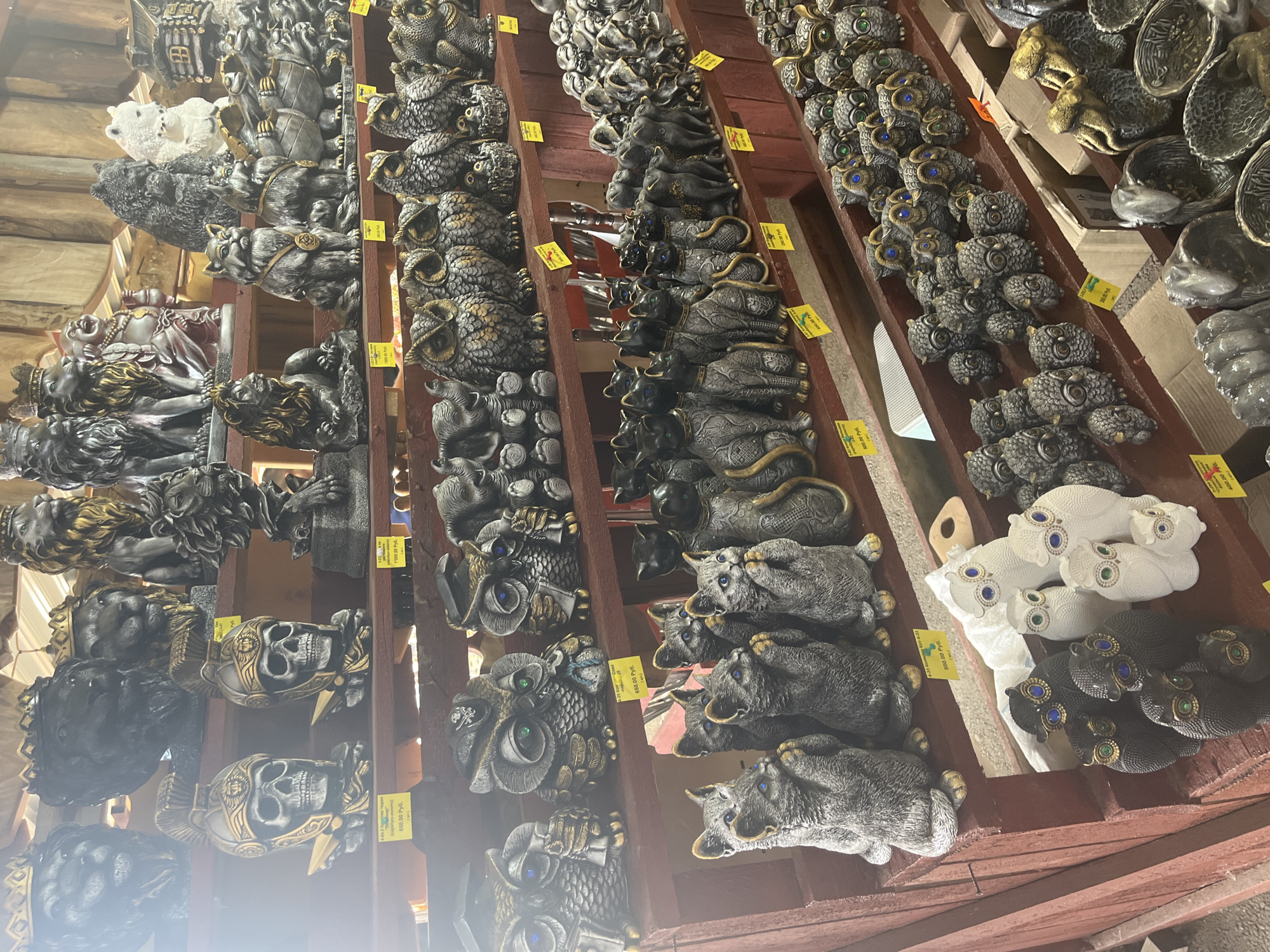 The image shows a market stall in Plastunovskaya, Russia, filled with an assortment of decorative figurines. The stall is organized with multiple shelves displaying various items. The top shelf features larger figurines, including lion heads with crowns, a seated lion, and a Buddha-like figure. Below that, there are skull-shaped figurines with Roman-style helmets, followed by a collection of owl figurines in different sizes and colors. The lower shelves are populated with numerous cat figurines in various poses and styles, as well as more owl figurines, and other animal figures such as bears and turtles. The figurines appear to be made of stone or a similar material, with intricate details. Some items have price tags in Russian rubles. The stall has a rustic wooden structure. There are no people visible in the frame.