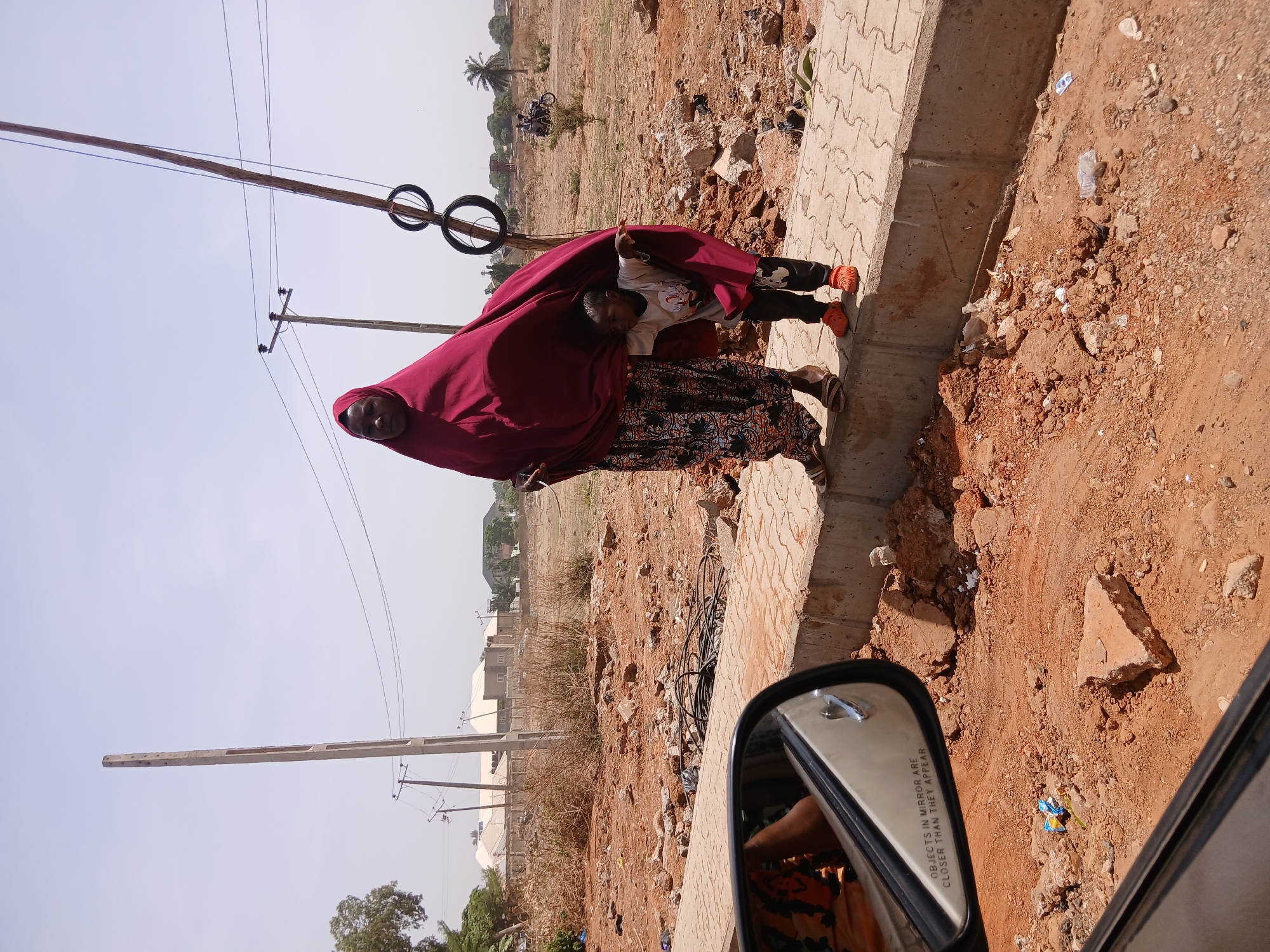 A medium shot, taken from inside a car, captures a woman in Unguwar Shanu, Nigeria. She is wearing a long, dark red hijab covering her head and most of her body, and a long, patterned skirt. A small child is partially concealed under her hijab, wearing a light-colored shirt and dark pants, with orange shoes.
They are standing on a concrete curb bordering a red, dirt lot. Overhead, utility poles and wires are visible against a light sky. In the background are low buildings. The side mirror of the car is visible in the lower right corner, with the inscription "OBJECTS IN MIRROR ARE CLOSER THAN THEY APPEAR". Debris litters the ground beside the curb.