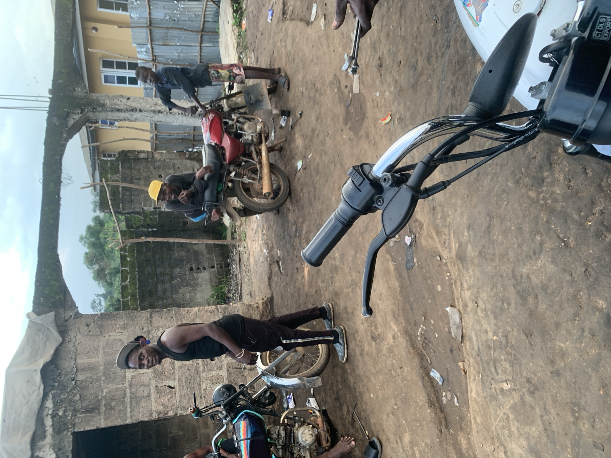 The image depicts a group of individuals engaged in activities related to motorcycles in what appears to be an outdoor workshop or repair area in Shagamu, Nigeria. The environment is characterized by a dirt or gravel ground with scattered debris, including pieces of plastic and what looks like discarded packaging. In the background, there are rudimentary structures constructed from concrete blocks and corrugated metal fencing, suggesting a developing urban or semi-urban setting.
Several people are visible. On the left, a man wearing a black tank top and dark pants stands next to a motorcycle. He has a light-colored cap tilted on his head and is looking towards the camera. Another motorcycle, a red one, is in the middle ground. Two men are associated with it; one, wearing a yellow cap, is seated on the motorcycle and appears to be in conversation or observing something. The second man, wearing a dark shirt and patterned shorts, is standing near the red motorcycle, holding onto its handlebars. In the foreground, a motorcycle's handlebars and part of its front are visible, implying the photo is taken from the perspective of another motorcycle.
The overall scene suggests a functional, informal setting where motorcycle repair or maintenance is taking place, common in many Nigerian cities like Shagamu. The lighting indicates it is daytime, possibly late afternoon or early morning, given the soft quality of light and the slight overcast sky.