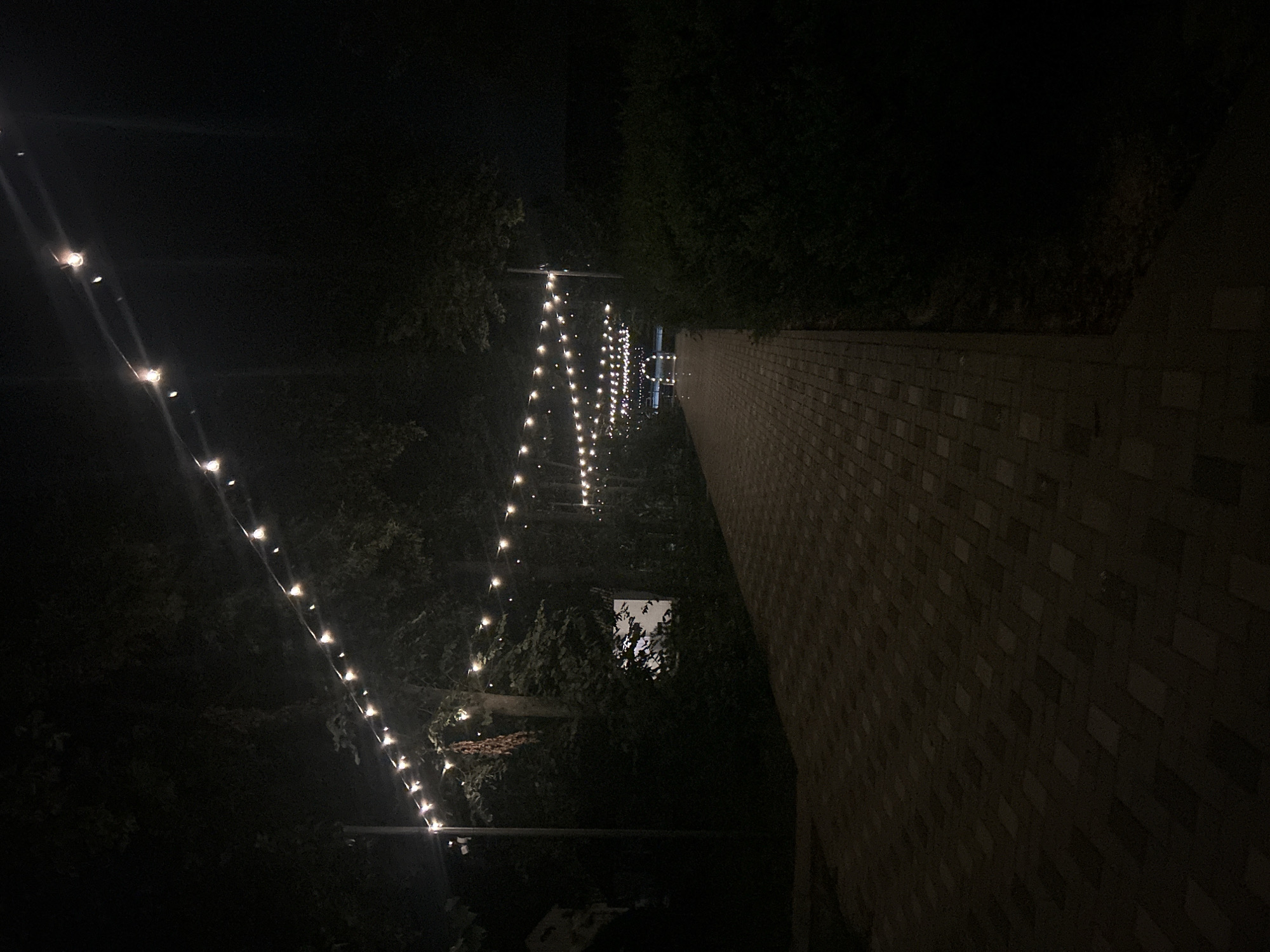 The image depicts a pathway at night, illuminated by strings of lights. The pathway, made of brick pavers, is bordered by dark foliage and trees. In the distance, the pathway leads towards an illuminated archway. The scene appears to be an outdoor setting, possibly a park or event venue, in Kudashivka, Ukraine. No people are visible in the image.