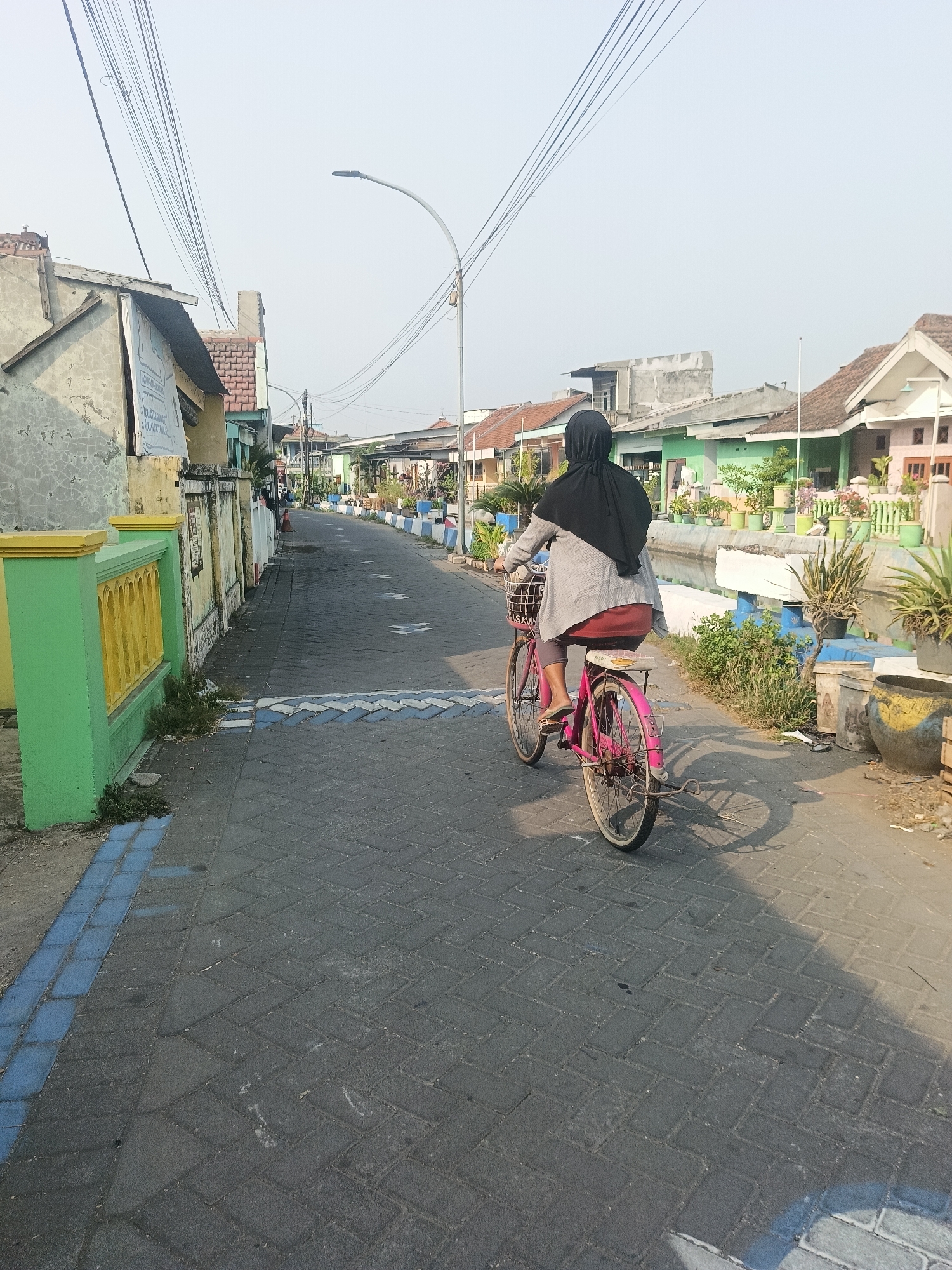 A woman wearing a black hijab and a grey cardigan is riding a pink bicycle down a paved street in Gedangan, Indonesia. The street is lined with houses on both sides, some of which have colorful fences and potted plants. Overhead, a tangle of electrical wires crisscrosses the sky, with a streetlamp visible in the distance. The sun is casting long shadows, suggesting it is either early morning or late afternoon. The bicycle has a basket in the front and a rack in the back. The woman is wearing sandals. The pavement is made of interlocking brick pavers. There are some water channels alongside the road.