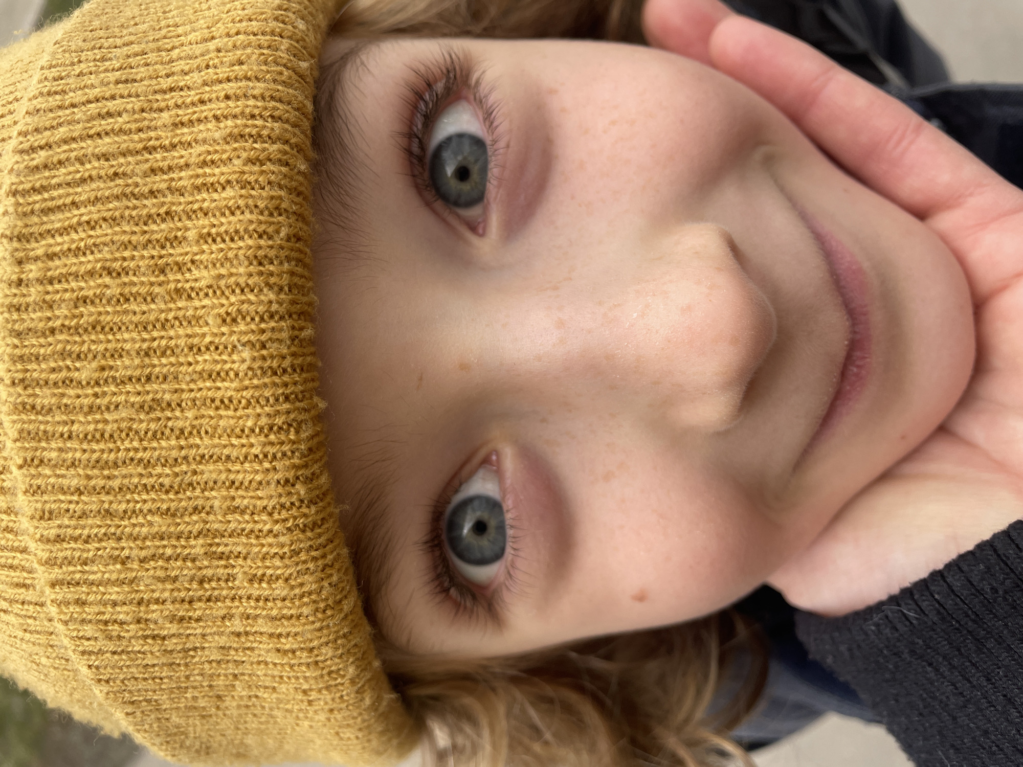 A close-up, off-kilter portrait of a young person is shown. The subject's face fills most of the frame. They have light skin with freckles, light-colored eyes, and wear a yellow knit cap. The subject is positioned with their head tilted to the right. A hand is gently holding the subject's face near their mouth. A small portion of a black garment can be seen near the right edge of the frame. The background is out of focus and indeterminate. There is no information to suggest this picture was taken in the city of Moscow, Russia.