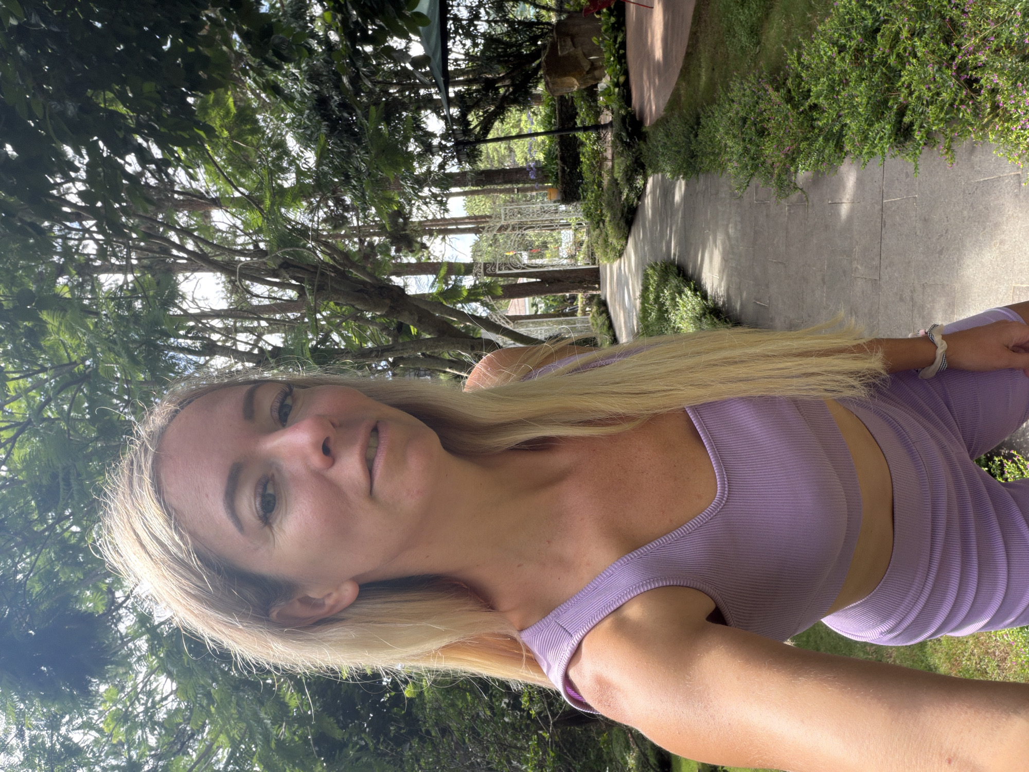 In the city of Xuân Trường - Đà Lạt, Vietnam, a blonde woman wearing a lavender athletic set is taking a selfie outdoors. She stands on a paved pathway lined with lush green bushes and trees. The pathway curves to the right, leading towards a white ornate gate structure in the background, with more trees and foliage. Sunlight filters through the leaves, casting dappled shadows on the ground. To the right of the pathway, there is an elevated red seating area with a green umbrella and a large rock. The woman's arm, extended for the selfie, is in the foreground on the left, and her hair cascades over her shoulder.