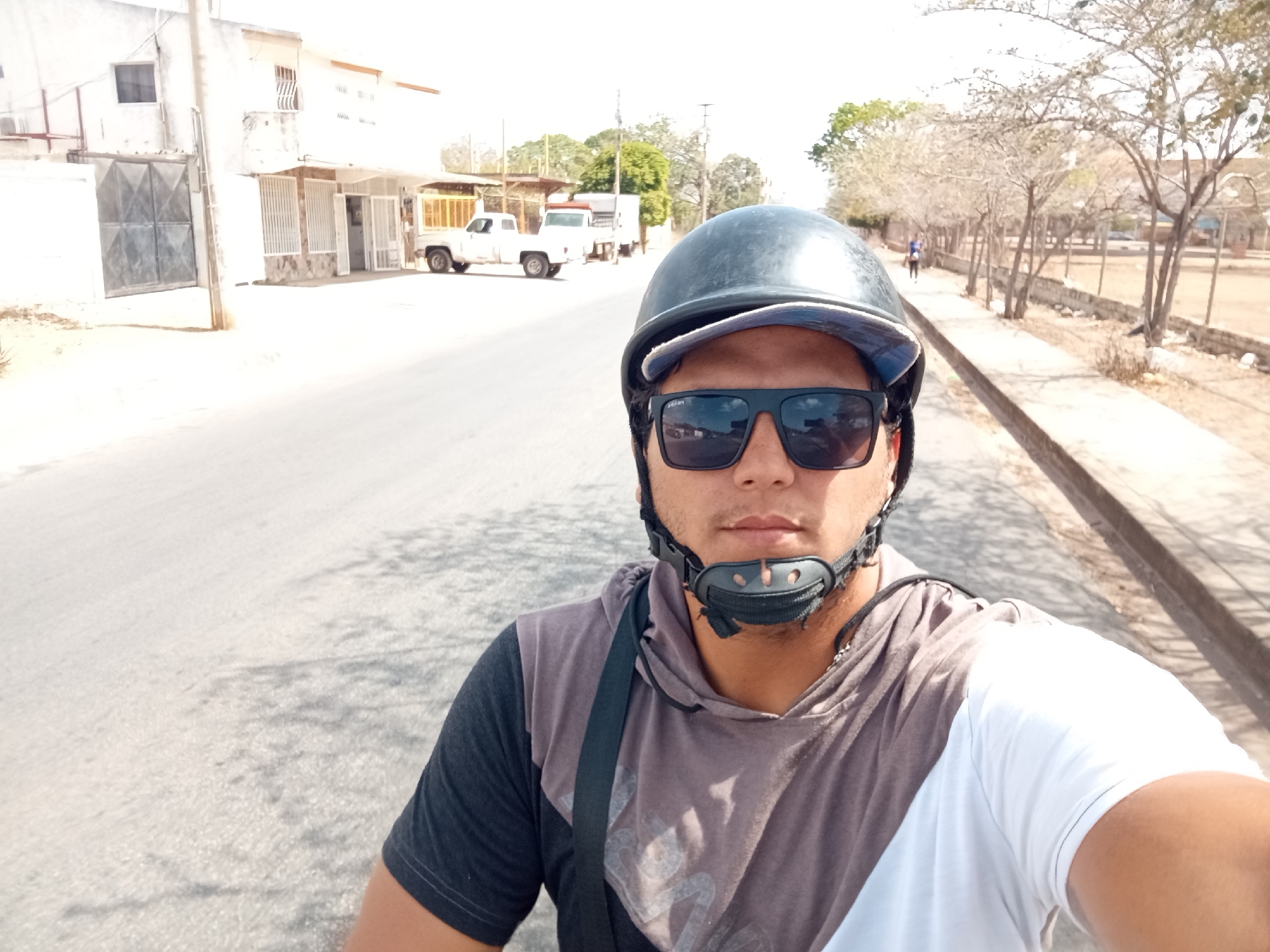 A medium shot depicts a man wearing a dark helmet, sunglasses, and a multi-toned hoodie. He's on a paved street in Juan Griego, Venezuela. In the background, there are white buildings, parked white trucks, and trees lining the sidewalk. A person is walking in the distance on the sidewalk.