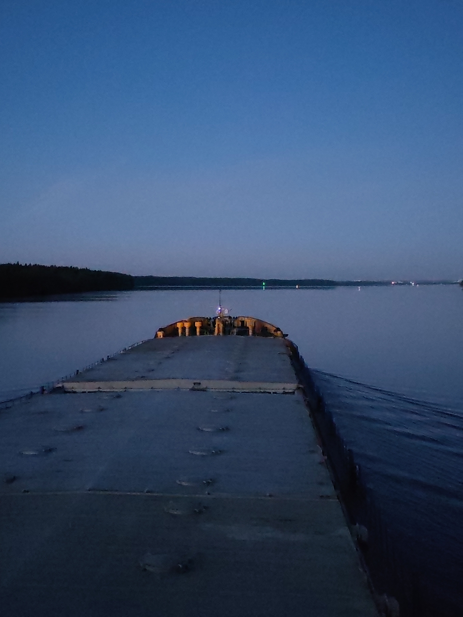 The image depicts a stern view from a large, flat-decked vessel navigating a wide body of water during twilight, near Yakunina Gora, Russia. The dark grey deck of the vessel extends from the foreground towards the center, featuring visible panel lines and several circular fixtures. Toward the far end of the deck, a cluster of illuminated yellow, cylindrical structures and a mast emitting a blue light are visible. A wake trails from the vessel's starboard side. A dense, dark forested shoreline forms the left bank of the waterway, receding into the distance. Scattered across the distant water, multiple lights are visible, including a prominent green light on the left and several fainter reddish-orange lights further to the right, indicative of navigation markers or distant shore features. The sky above is a deep, clear blue, transitioning subtly towards the horizon, consistent with low light conditions. No people are visible.