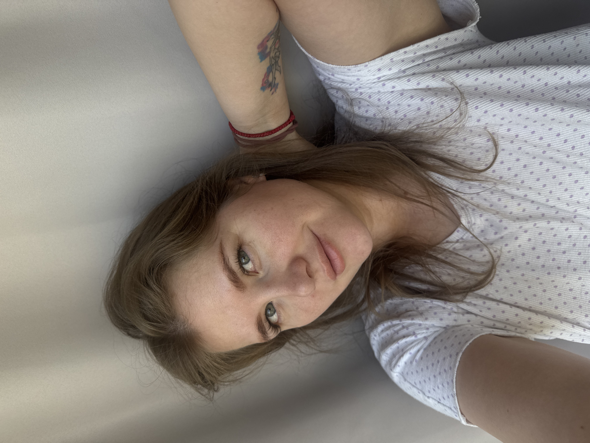 A young woman with fair skin and light brown hair is lying down, posing for a close-up selfie. She has green eyes and freckles on her face. She's wearing a white pajama top with light purple polka dots. On her left arm, she has a tattoo in multiple colors and two red string bracelets. The background is a plain gray surface. The photo appears to have been taken indoors. There is no information to determine if the photo was taken in Ферма, Russia.