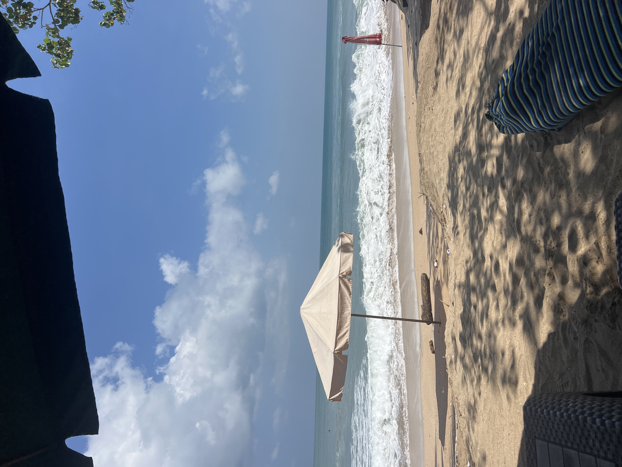 The image depicts a serene beach scene in Kuta, Indonesia. A large, cream-colored umbrella stands on the sand, providing shade. In the background, the ocean churns with white-capped waves, meeting a sandy shore. A red umbrella is visible further down the beach. The foreground shows a beach chair and a striped towel, suggesting a relaxing day at the beach. The sky is a brilliant blue, dotted with fluffy white clouds. The overall impression is one of tropical tranquility and a perfect day for beachgoers.