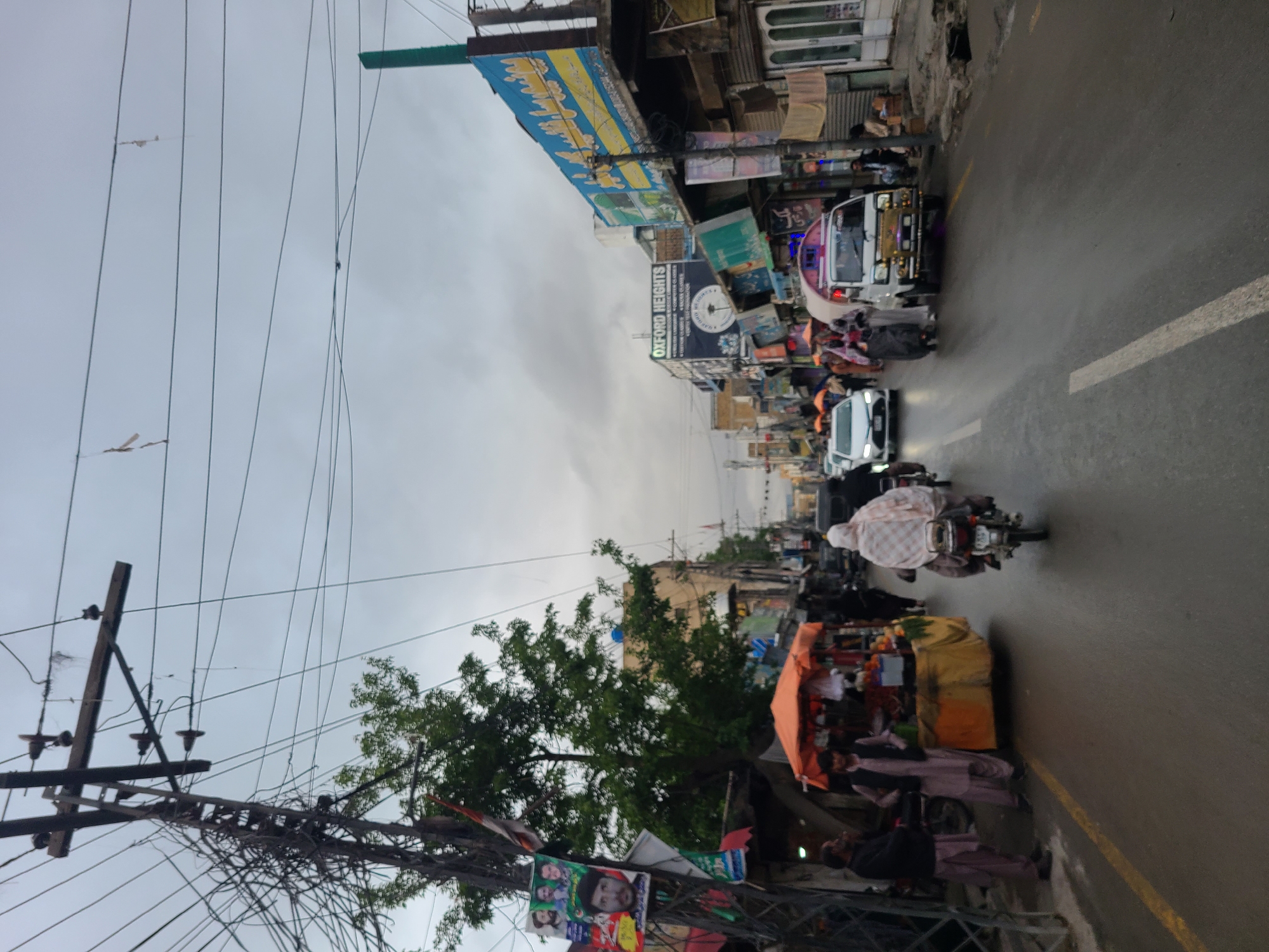 The image depicts a street scene in Quetta City Tehsil, Pakistan. The street is wet and lined with buildings adorned with signage. There is a variety of vehicles on the road, including an auto rickshaw, a white car, and motorcycles. Pedestrians and vendors are present alongside the road. Overhead, there are utility poles with tangled wires. The sky is overcast.