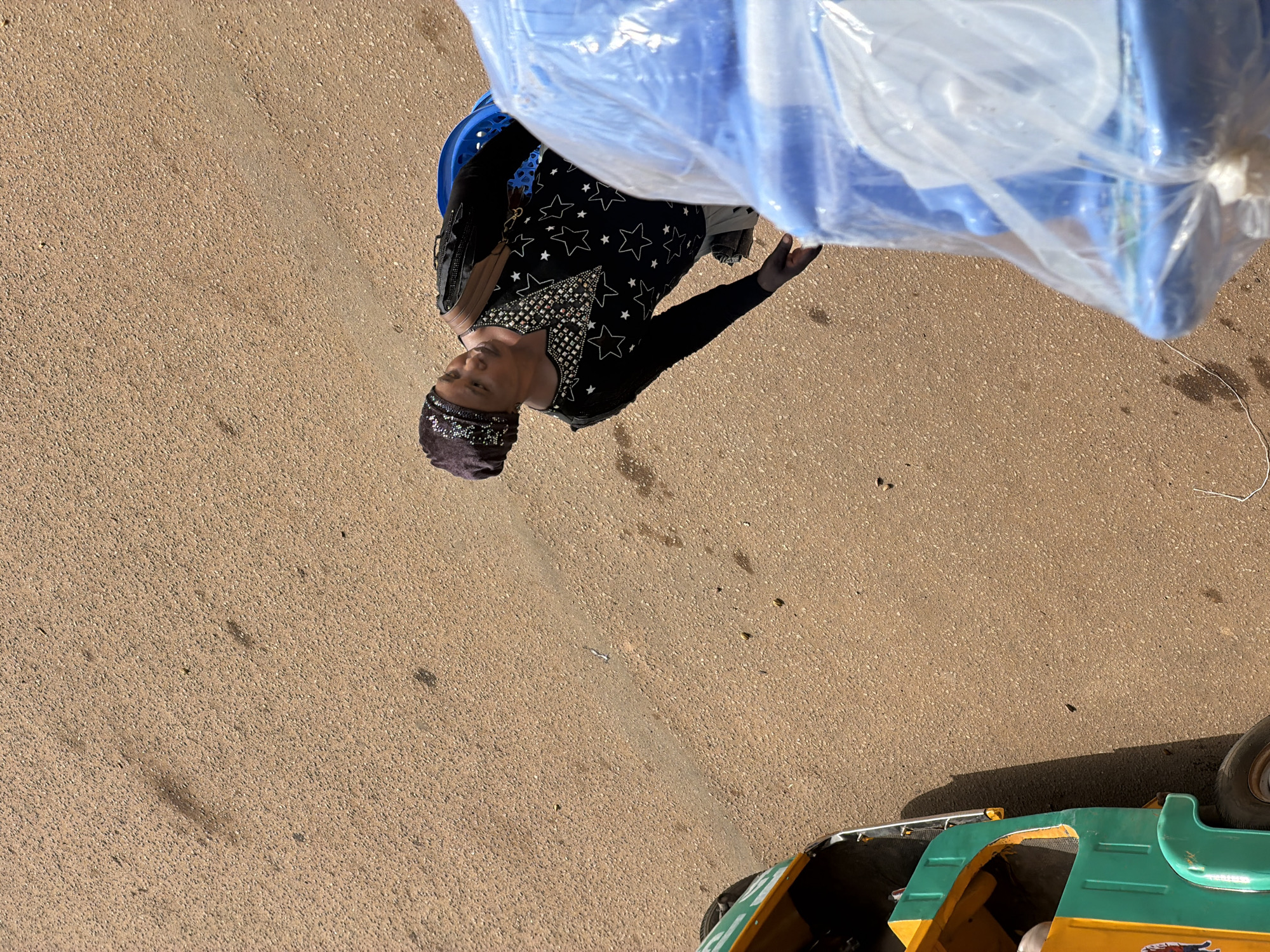 In Bukuru, Nigeria, a woman wearing a dark headscarf and a black, star-patterned shirt stands on a dirt road. She is looking to her right and has a brown strap across her shoulder, suggesting she is carrying a bag. In the foreground, a bright blue plastic object wrapped in clear plastic is partially visible, positioned in front of the woman. To the left of the frame, the front section of a green and yellow autorickshaw is visible, its wheel turned slightly. The ground is a rough, sandy-brown texture, indicative of a typical road surface in the area, with scattered small debris and a faint shadow cast by the autorickshaw.