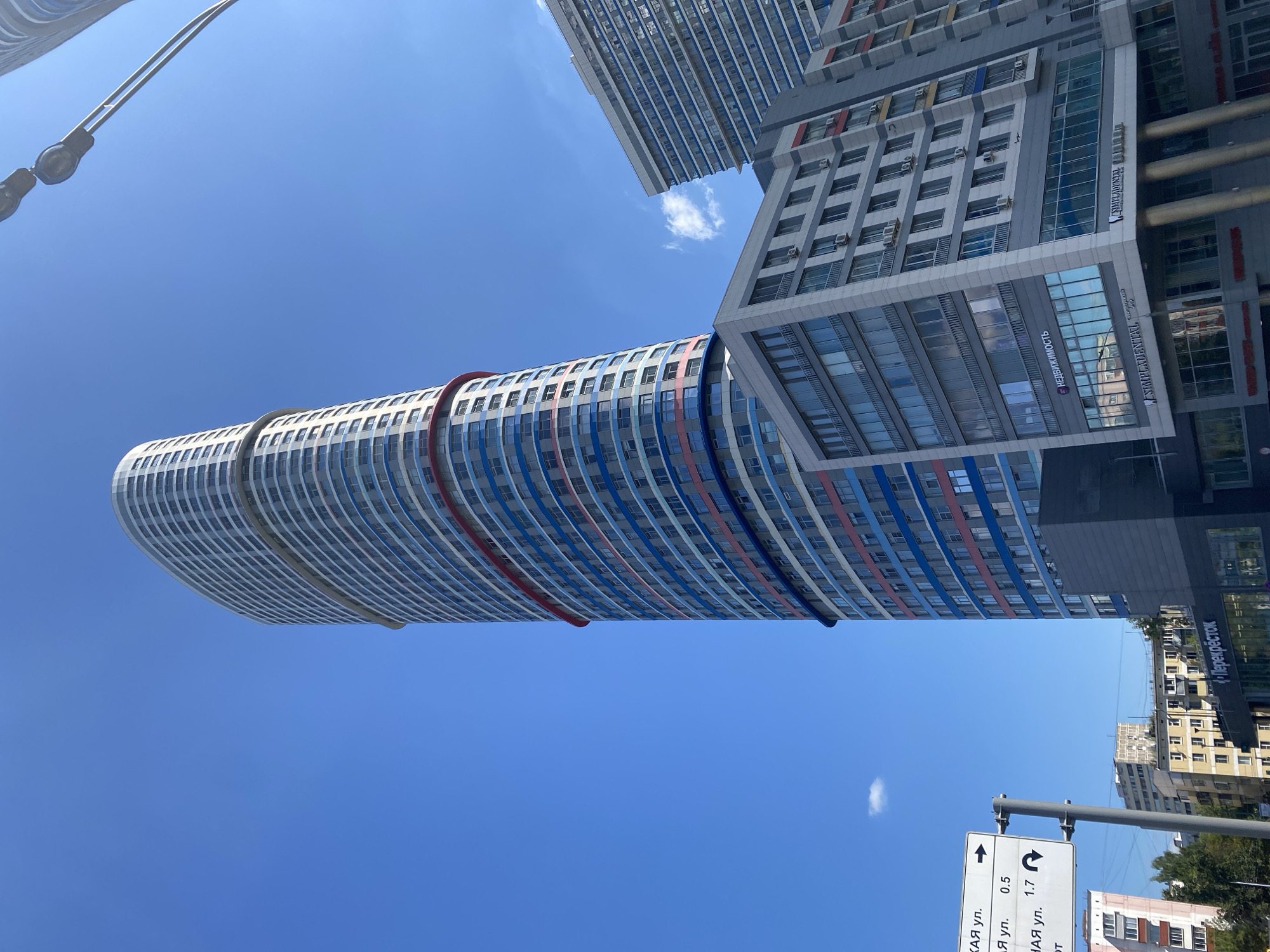 An eye-level outdoor shot captures a modern, cylindrical skyscraper against a clear blue sky. The skyscraper is predominantly white with horizontal bands of red, blue, and grey. A street sign with Cyrillic text and arrows indicating distances and directions stands to the left of the frame. In the foreground, a lower, grey building with large windows displays storefronts, including one with the text "НЕДВИЖИМОСТЬ" (real estate) and another with "SIMPLADENTAL". To the right, another tall building with a blue and white facade is partially visible. The scene appears to be in an urban environment with trees and other buildings in the background.