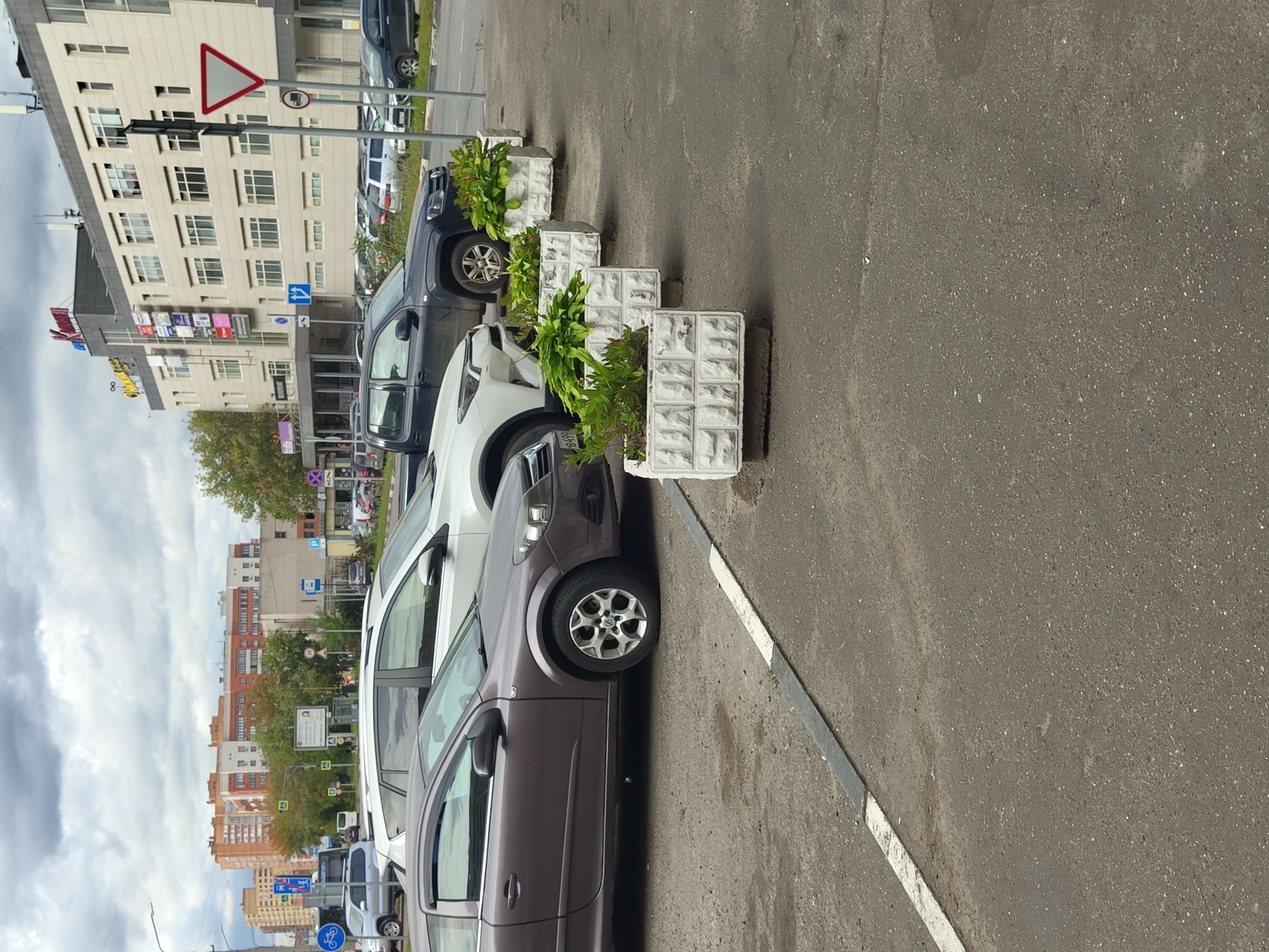 The image shows a row of cars parked along a street in Zhukovsky, Russia. The cars are parked next to a sidewalk with several white planters containing green plants. In the background, there are buildings, trees, and a cloudy sky. The street is paved with asphalt and has a white line marking the parking spaces. The time of day appears to be daytime.