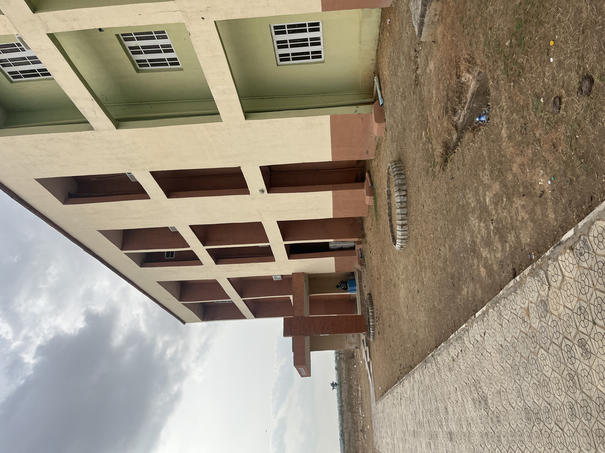 The image shows a multi-story building with light-colored walls and brown trim in Baganakwo, Nigeria. The building has rows of rectangular windows with white frames. The ground surrounding the building is mostly bare earth with scattered debris. There's a patterned concrete walkway adjacent to the building. The sky is overcast with gray clouds. There is a circular stone structure on the ground near the building.