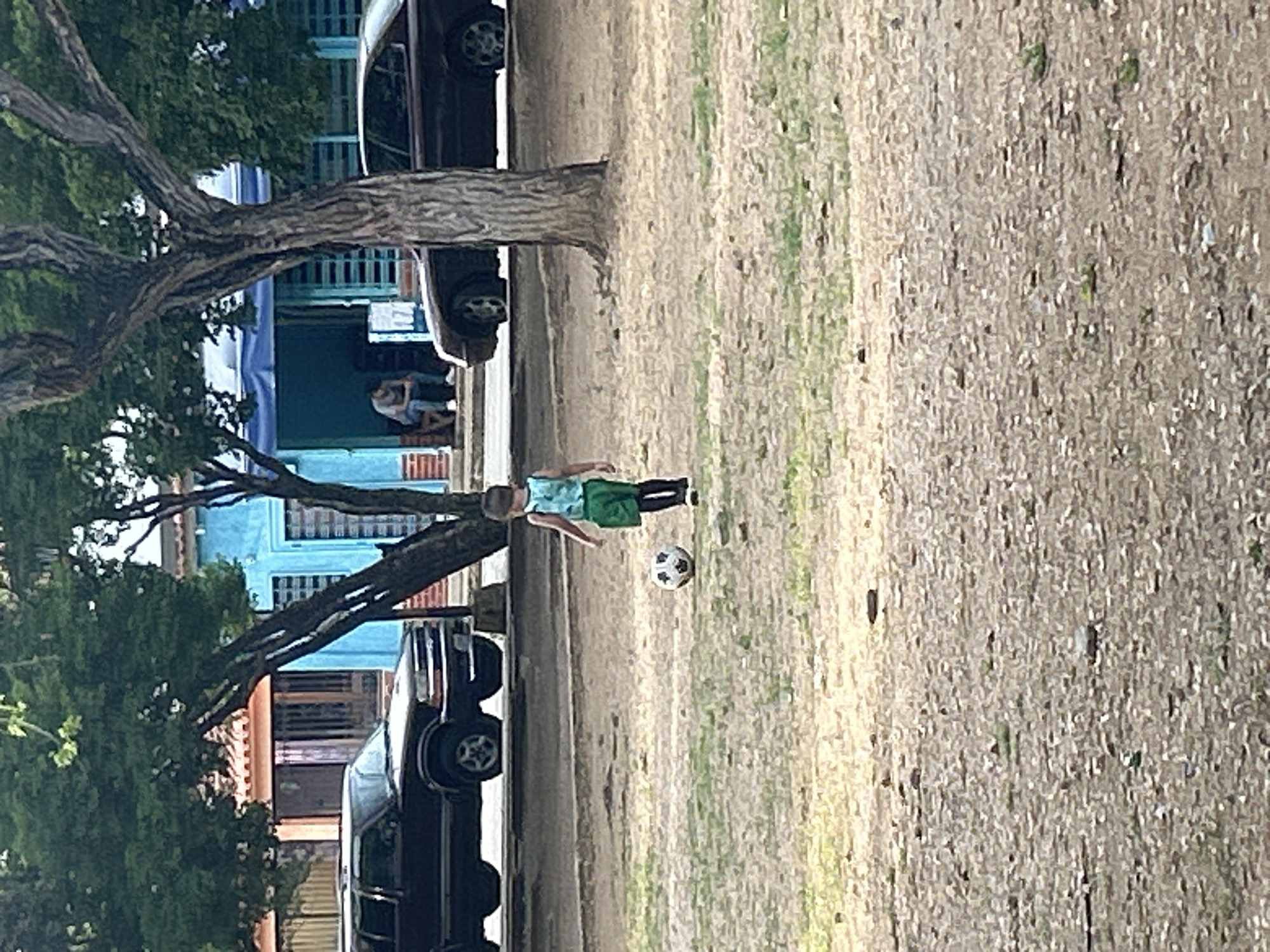 In Santa Ana, Venezuela, a young boy is playing with a soccer ball in a park area. He is wearing a teal tank top, green shorts, and black leggings. The boy is standing in front of a soccer ball. In the background, there are trees, cars, and buildings. One building is painted blue and has a blue awning. A person is sitting on a bench in front of the blue building. The ground is covered in dirt and some patches of grass.