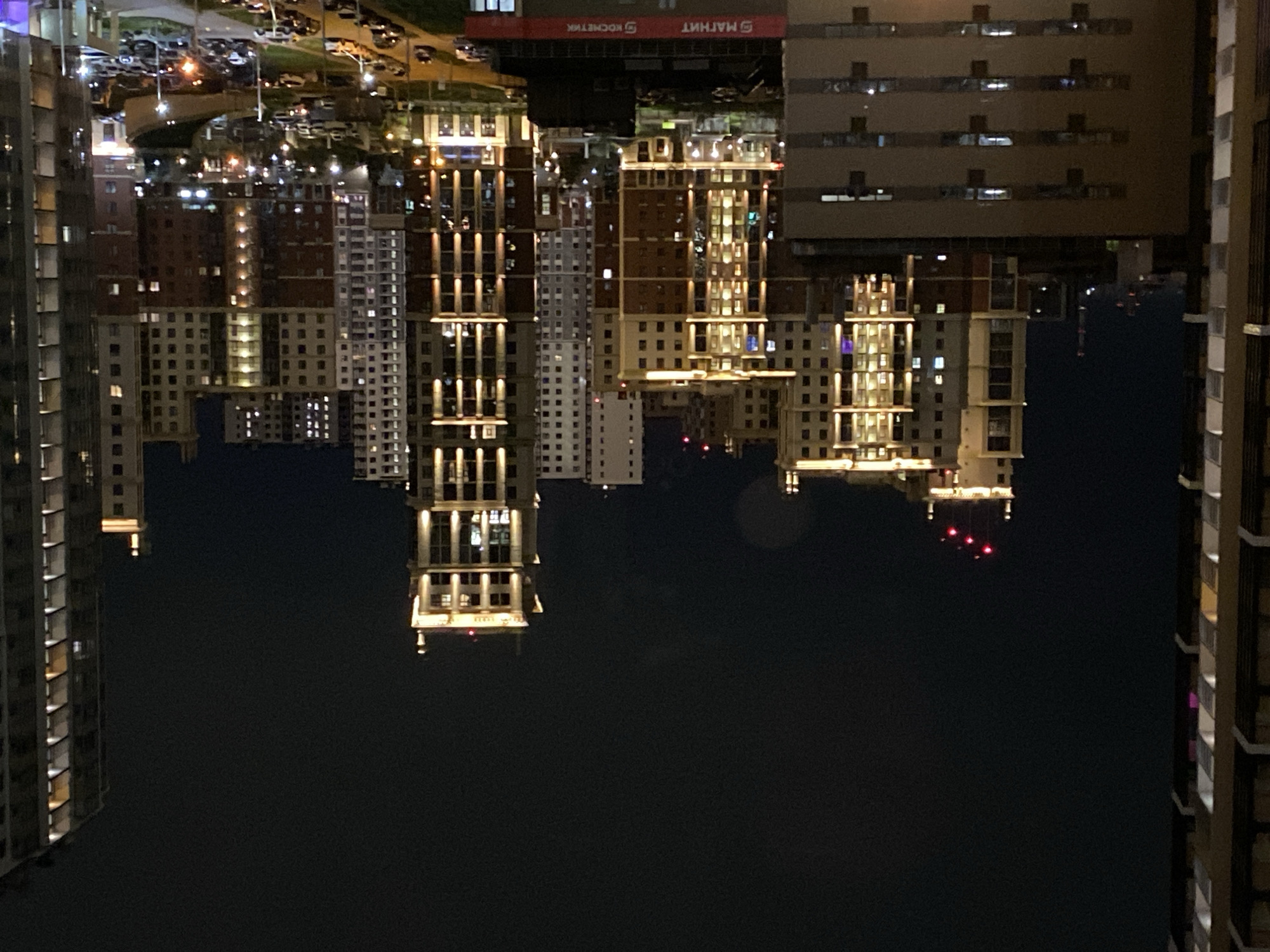 The image depicts an upside-down view of illuminated buildings at night. Streetlights and parked cars are visible near the tops of buildings. Building signs are visible. The overall setting suggests an urban environment, with the buildings being residential or commercial. The lighting is predominantly artificial from windows and streetlights. The background is dark, indicating a nighttime setting.