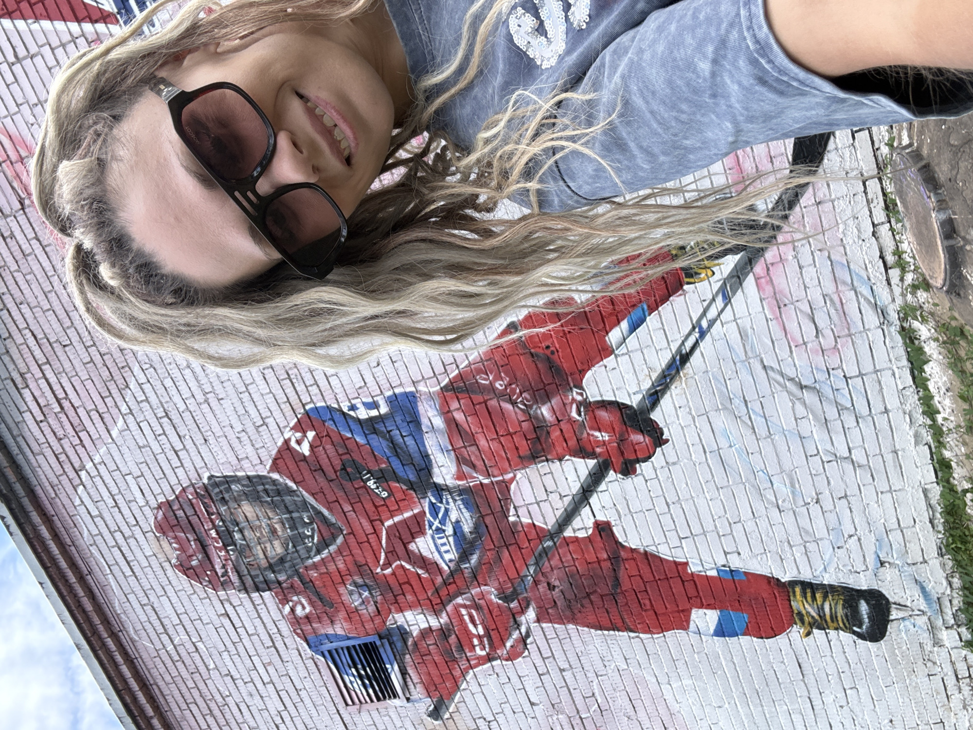 In Rusanovo, Russia, a young woman with long, blonde, braided hair and dark sunglasses poses for a selfie in front of a brick wall mural. The mural depicts a hockey player in a red and blue uniform, holding a hockey stick. The woman is wearing a grey t-shirt. The sky is visible in the background.