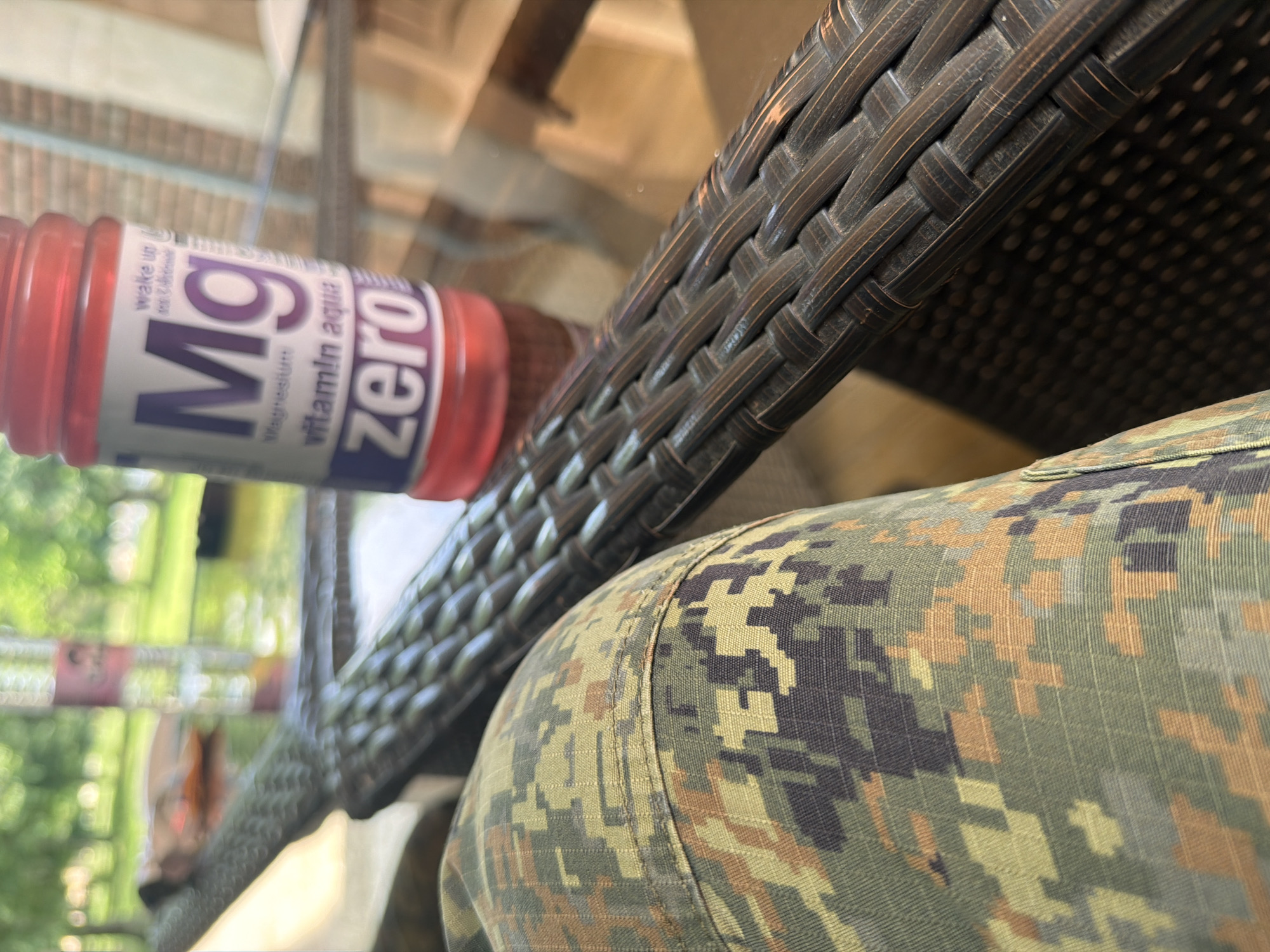 The image shows a close-up of a wicker chair, a bottle of "Mg vitamin aqua ZERO" drink, and a person wearing a camouflage-patterned garment, possibly a jacket or shirt. The drink bottle is partially visible and positioned next to the chair. The setting appears to be outdoors, with blurred greenery visible in the background, suggestive of a garden or patio. There is no information in the image to identify the specific location as Dolna Mitropolia, Bulgaria.