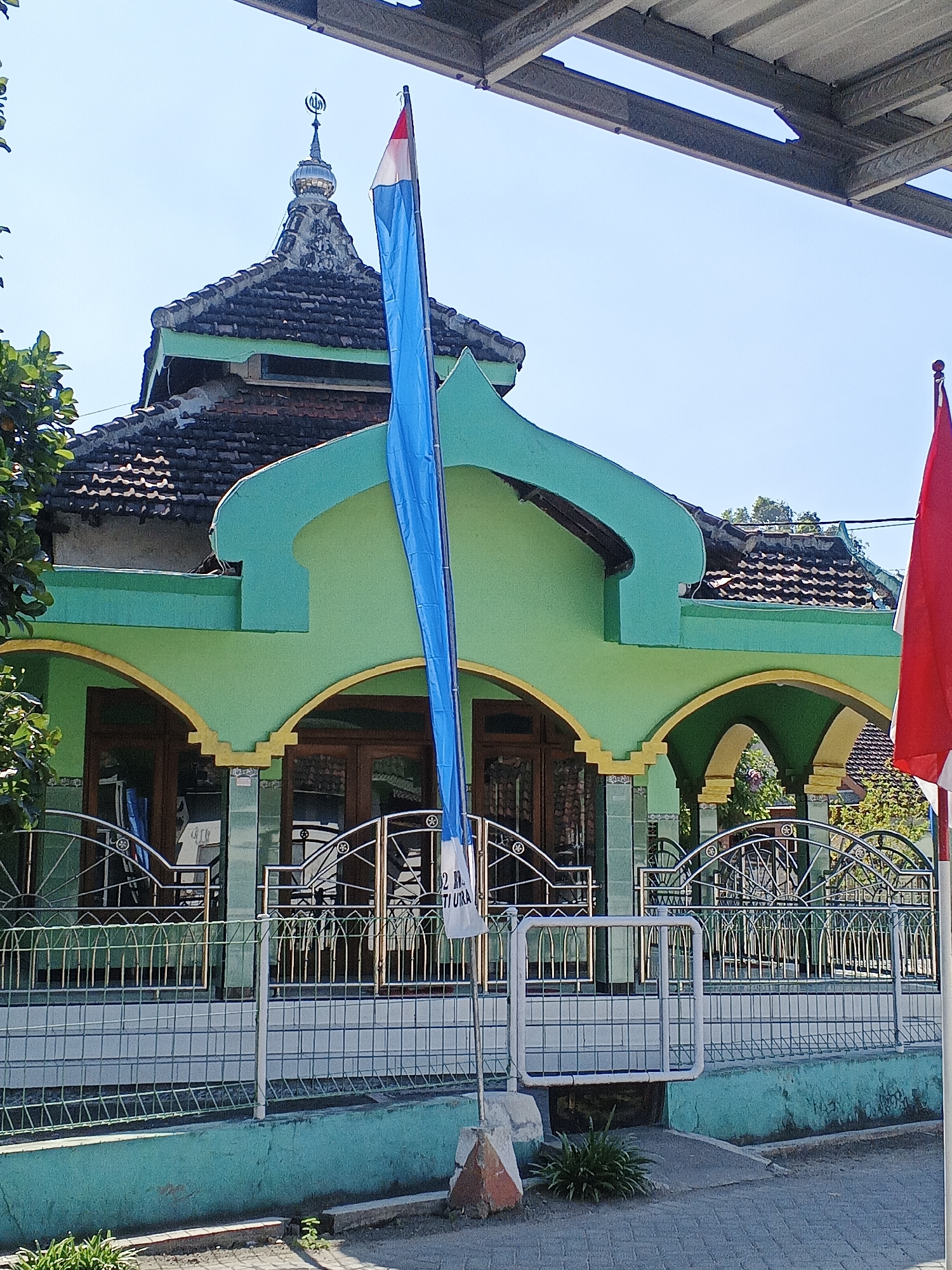 The image depicts the exterior of a mosque in Kemangsen, Indonesia. The mosque has a green facade with arched entrances and a tiled roof. A minaret with a crescent moon finial rises from the roof. Two flags are prominently displayed in front of the mosque: a blue and white flag and a red flag. A metal fence surrounds the mosque's courtyard. The sky is clear and blue, indicating fair weather. The overall scene is one of a religious building under a bright, sunny day in a presumably quiet, possibly rural, setting.