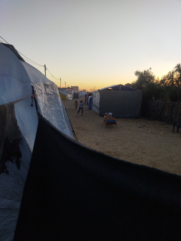 The scene depicts a makeshift encampment in Khan Yunis, Palestinian Territory, at either dawn or dusk, characterized by a sky gradient from light grey overhead to orange-yellow at the horizon. The ground throughout is exposed light brown sand or dirt. In the immediate foreground, a large, light-colored tent-like structure, likely constructed from tarp, occupies the left side, partially obscured by a prominent black fabric element in the lower left. Dark, tattered fabric or netting hangs from the side of this tent.
A dirt path extends through the midground, flanked by multiple temporary shelters of various sizes and materials, including tarps and fabric, forming rows that recede into the background. A distinct dark grey or green makeshift structure with a visible roof is present on the right side of the path. In front of this structure, a small, low table with a red base and dark top holds several colorful, indistinct objects. A dark, partial object, possibly a chair, is visible at the far right edge.
Two young children, wearing light and dark clothing, are observed actively running from left to right across the sandy path in the midground. Overhead, electrical wires are strung between utility poles visible above the shelters. The far right background features a cluster of dark-foliaged trees and bushes silhouetted against the horizon. Distant urban structures are faintly outlined against the transitional sky in the far background.