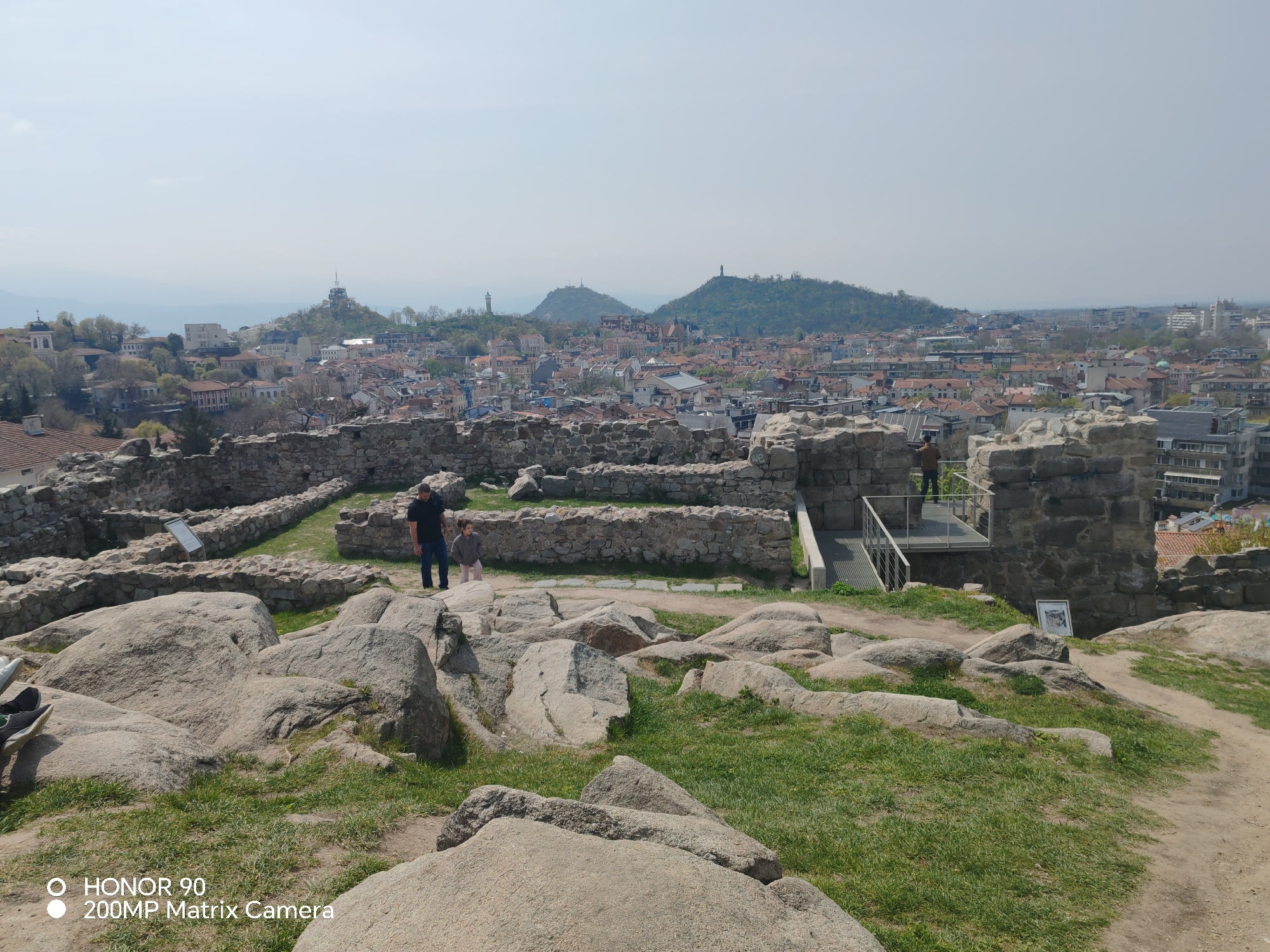 The image shows a view of Plovdiv, Bulgaria, from the Nebet Tepe archaeological site. Nebet Tepe features ancient ruins, visible as stone walls and foundations scattered across the rocky terrain in the foreground. Some grassy areas grow around the rocks. A path runs through the rock formations.
In the middle ground, there's a father with his child. Next to the ruins on the right, there's a modern bridge.
The background reveals a dense urban landscape of Plovdiv, characterized by red-tiled roofs and various buildings. Three of the hills surrounding Plovdiv stand out: Bunardzhik Hill, Dzhambaz Tepe, and Taksim Tepe. The sky is overcast and hazy.
A smartphone watermark is visible in the lower left corner.