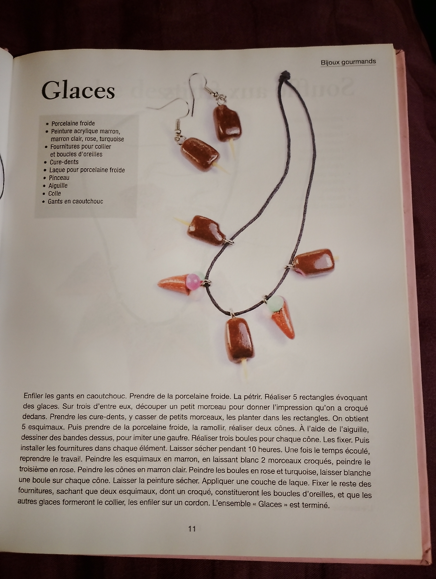 The image is a close-up, high-angle shot of an open book, displaying a page with text and an accompanying illustration. The page is titled "Glaces" (meaning "Ice Creams" in French) and also has a smaller heading "Bijoux gourmands" (meaning "Gourmet Jewelry").
The illustration features a necklace and a pair of earrings, both designed to resemble ice cream cones and popsicles. The jewelry is made from a brown material, likely clay or polymer clay, representing chocolate ice cream. Some pieces are shaped like elongated rectangles on sticks (popsicles), while others are conical (ice cream cones). The ice cream cones have a pinkish-orange cone part and a small pink or turquoise ball on top, representing a cherry or topping. The jewelry is arranged on a plain white background.
The text on the left side of the page lists the materials needed to create the jewelry, including "Porcelaine froide" (cold porcelain), acrylic paint in brown, light brown, rose, and turquoise, findings for necklaces and earrings, toothpicks, varnish for cold porcelain, a paintbrush, a needle, glue, and rubber gloves.
The text on the right side of the page provides instructions for making the jewelry. It describes how to work with the cold porcelain, shape it into rectangles and cones, and paint them to resemble ice cream. It also mentions the assembly of the jewelry, including attaching findings and stringing the pieces onto a cord for the necklace and hooks for the earrings.
The bottom of the page shows the number "11", indicating it is page 11 of the book. The overall aesthetic is a craft or DIY tutorial focused on creating food-themed jewelry. The book is lying on a textured, dark reddish-brown surface, possibly fabric.
