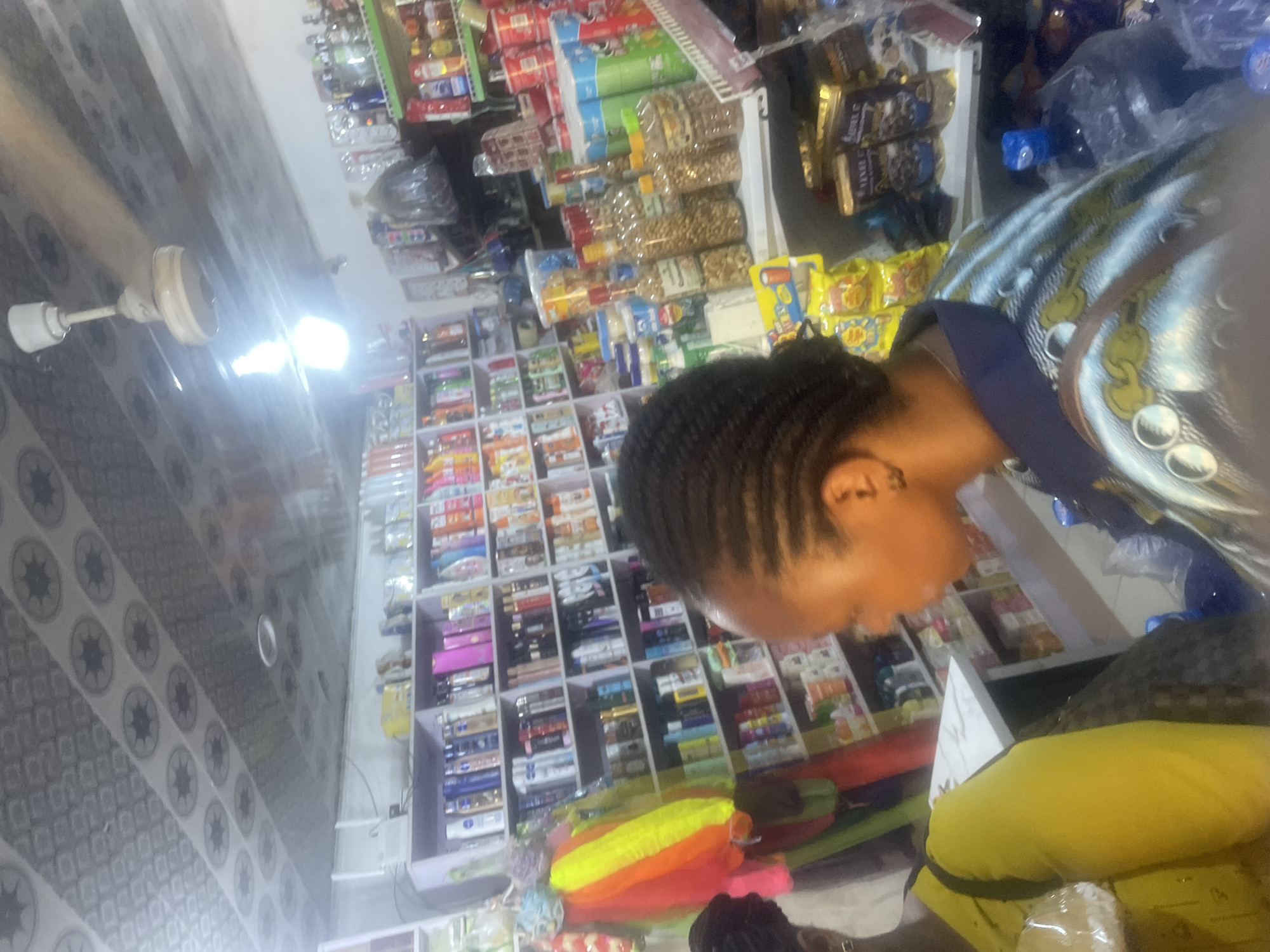 The image shows a retail store, likely a small convenience store or kiosk, with shelves stocked with various goods. The scene is indoors, with fluorescent lighting illuminating the products. The shelves are filled with packaged snacks, beverages, and other household items. In the foreground, a person, possibly a woman, is partially visible, with their hair styled in braids. They are wearing a patterned shirt. The overall impression is of a busy retail environment, with goods neatly arranged on display. The background shows more shelves and products, creating a sense of depth and abundance. The image is taken at an angle, possibly to capture the entirety of the store's offerings. There is no clear indication of the time of day or weather conditions. The location context provided suggests this scene is in Abuja, Nigeria.