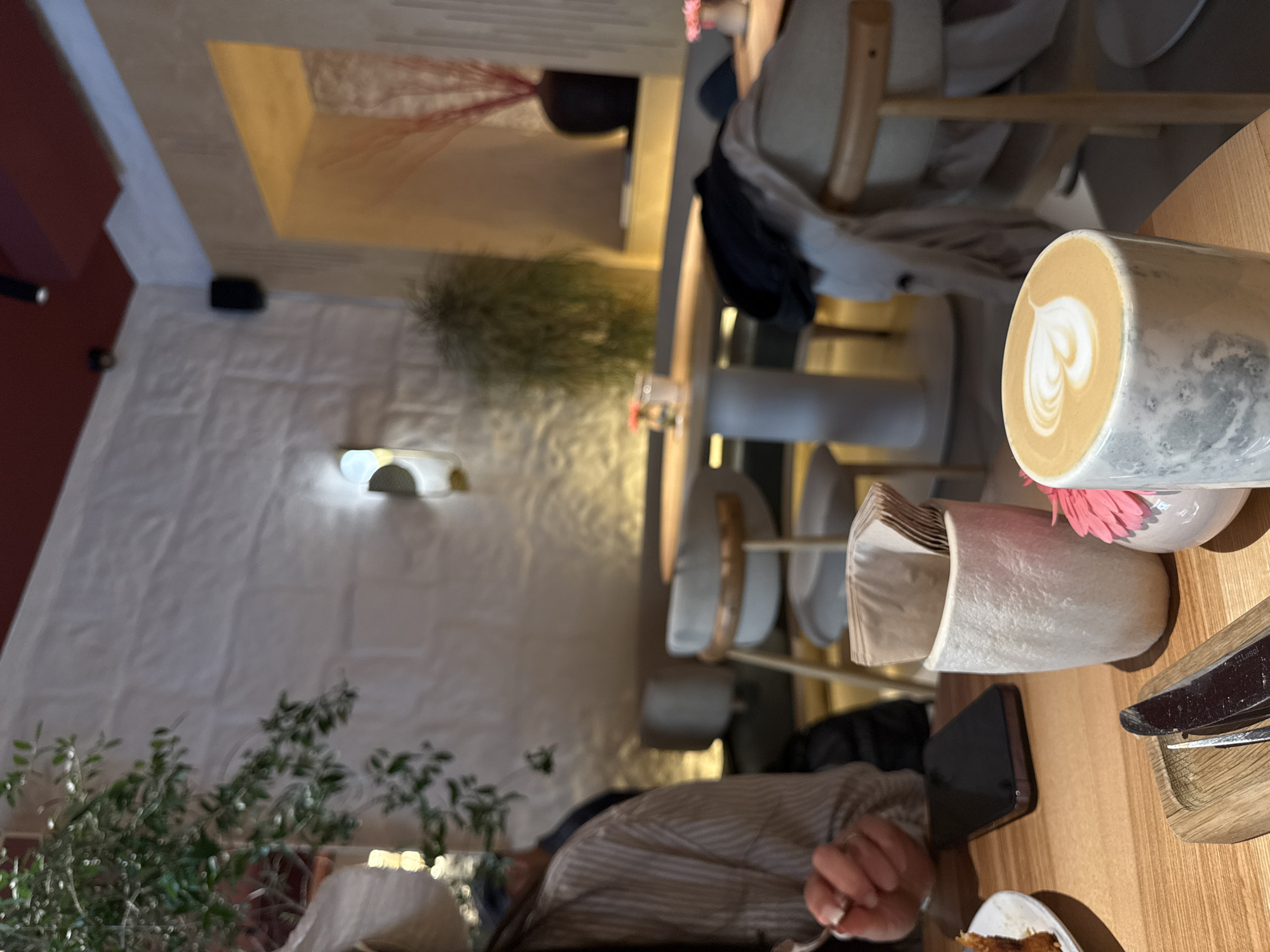 A slightly blurry, vertically oriented photo shows a table scene with a person at a restaurant, potentially in Yerevan, Armenia, given the user's prompt. In the foreground, a light wooden table is partly occupied by objects: a coffee mug with latte art, a holder containing napkins, a smartphone, a wooden plate with utensils, and a portion of food on a plate. A person wearing a striped shirt is seated at the table with a hand near the food.
The background displays a softly lit, textured white wall. A mounted, abstract light fixture is visible. In the distance, multiple chairs are positioned around tables, indicating a dining area. Plants are also visible in the background, adding to the restaurant's ambiance.