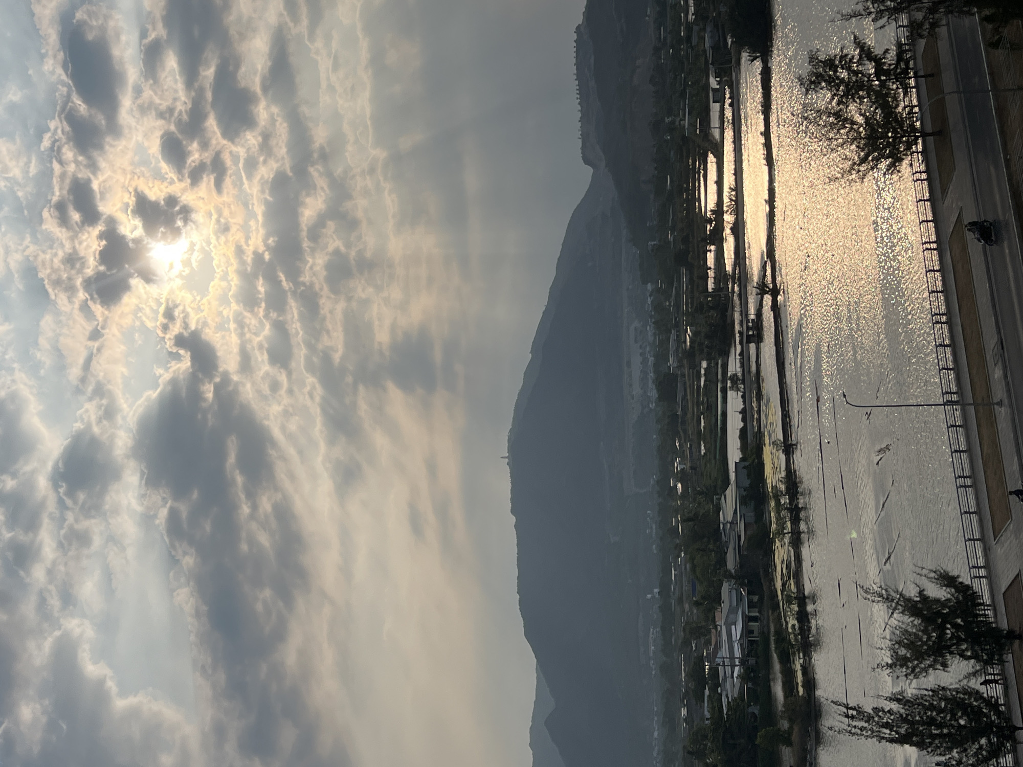 This wide-angle image depicts a scenic landscape, likely captured during sunrise or sunset, given the low sun position and the golden light illuminating the scene. The sky is a prominent feature, filled with dramatic, scattered clouds through which the sun actively breaks, casting bright, visible rays of light.
In the foreground and mid-ground, a tranquil body of water, possibly a river or bay, stretches across the frame. Its surface shimmers with intense golden reflections from the sun, creating a sparkling effect. On the immediate right bank of this water body, a paved road or pathway runs parallel, bordered by a black metal railing. Several dark, silhouetted trees, possibly coniferous, are visible along the path. A street light also stands near the road, and a dark shape that may be a parked motorcycle or scooter is discernible on the pavement near the railing.
Across the water, on the left bank, various buildings or houses are nestled at the base of a significant mountain range that forms the backdrop of the scene. The mountains appear hazy, suggesting atmospheric conditions or distance. A notable feature on one of the mountain peaks is a tall, slender structure, resembling a monument or statue, standing prominently against the sky.
The overall scene conveys a sense of peaceful natural beauty. Given the provided context, the location is the Nam Natural Heritage Area in Trang, Vietnam. No discernible text is visible within the image.