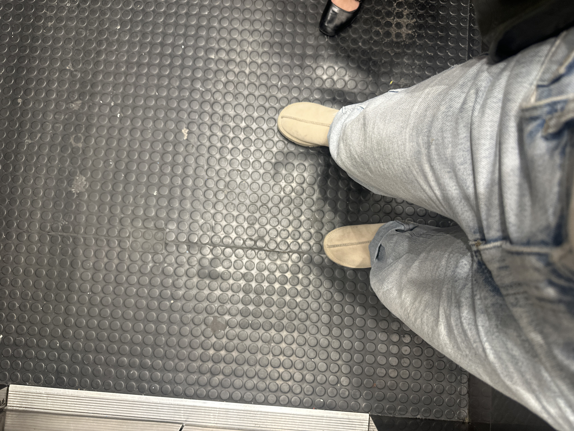 The image depicts a downward shot of a person standing on a black rubber-studded floor. The individual is wearing light-wash denim jeans and beige slippers. A portion of another person's black shoe is visible in the upper right corner of the frame. The floor has a metallic strip along the bottom edge. There is no information in the image to determine if this is in Mexico City.