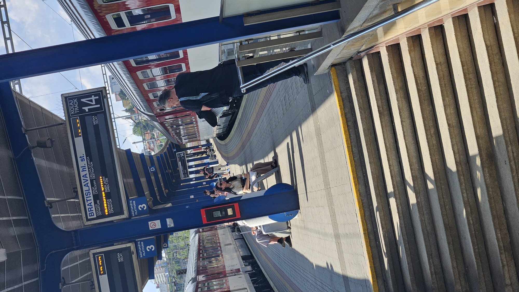 The image depicts a platform at Bratislava hl. st. (Bratislava Main Station) in Bratislava, Slovakia. A red and white train is parked at track 14. Several passengers are visible on the platform. In the foreground, a man in a black shirt and dark pants is standing, holding a jacket over his shoulder, looking towards the train. Further down the platform, a woman with sunglasses is seated on a bench, holding a bag. Another person is seated next to her with a suitcase. The platform is paved with concrete and has yellow markings along the edge. Overhead signs indicate train departures and platform numbers, with "PLATFORM 3" and "TRACK 14" clearly visible. The scene is illuminated by natural sunlight, casting shadows on the platform.