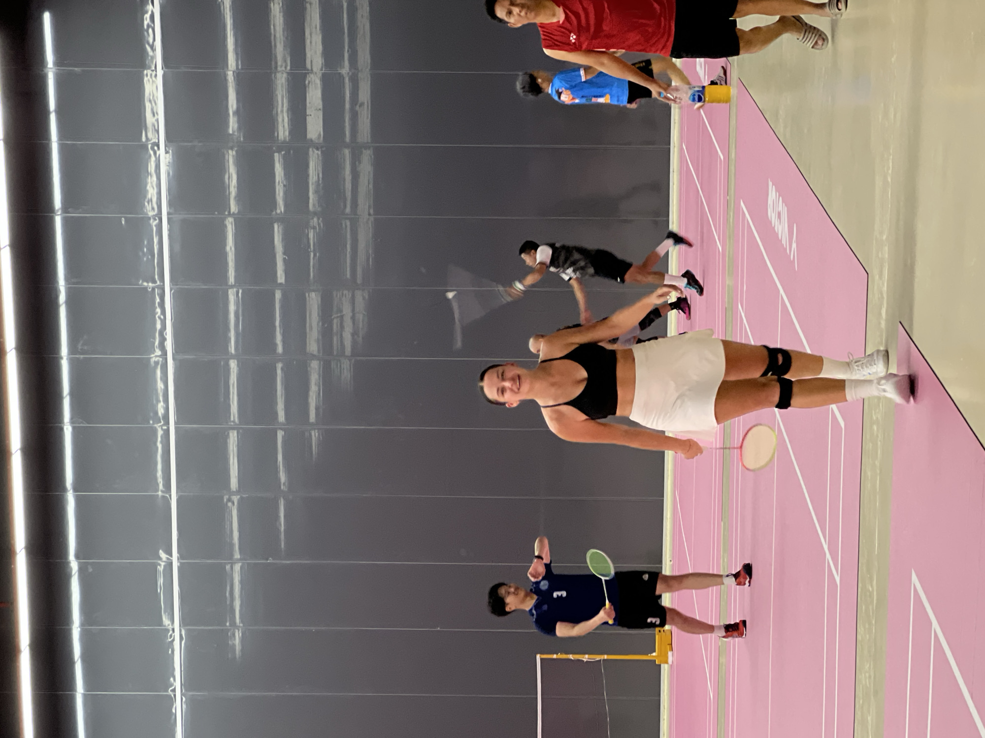 A group of people are playing badminton indoors on a pink court. In the foreground, a woman in a black sports bra and white skirt is looking at the camera and smiling, holding a yellow badminton racket. To her right, a man in a blue shirt and black shorts is in motion, ready to hit a shuttlecock. Further back, another man in a black and white shirt is also in action, and a man in a red shirt is standing. The walls are dark gray and tiled. The lighting suggests it is daytime, and the indoor setting indicates controlled conditions. The people appear focused and engaged in the game. Text is visible on the pink court, but it is not legible. The location is Ko Samui, Thailand.