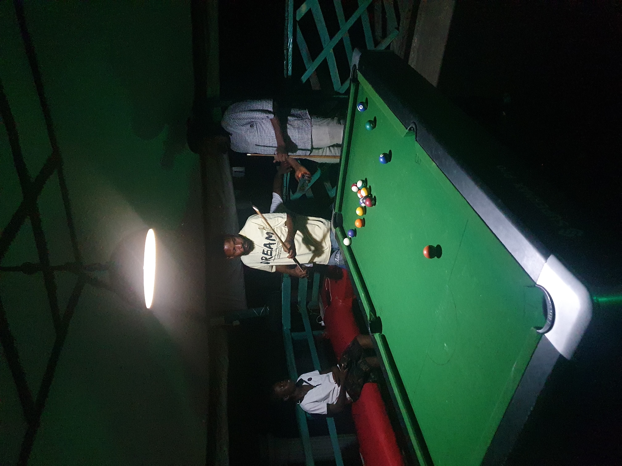 This is a medium shot of three men playing pool at night. The scene is outdoors, likely on a raised platform, with a green ceiling and dark green railings visible in the background. A bright, overhead light illuminates the pool table.
On the left, a young man in a white polo shirt and checkered shorts sits on a red bench, watching the game. In the center, a man in a cream-colored t-shirt with the word "DREAM" printed on it stands holding a pool cue, poised to take a shot. To the right, another man in a plaid shirt and light-colored pants stands with a pool cue, observing the game.
The pool table is green with a full rack of billiard balls and several balls scattered on the table. The environment suggests a casual, recreational setting. The image is consistent with a location like the rooftop bar at the Confluence Beach Hotel in Lokoja, Nigeria, known for its scenic views and recreational facilities.