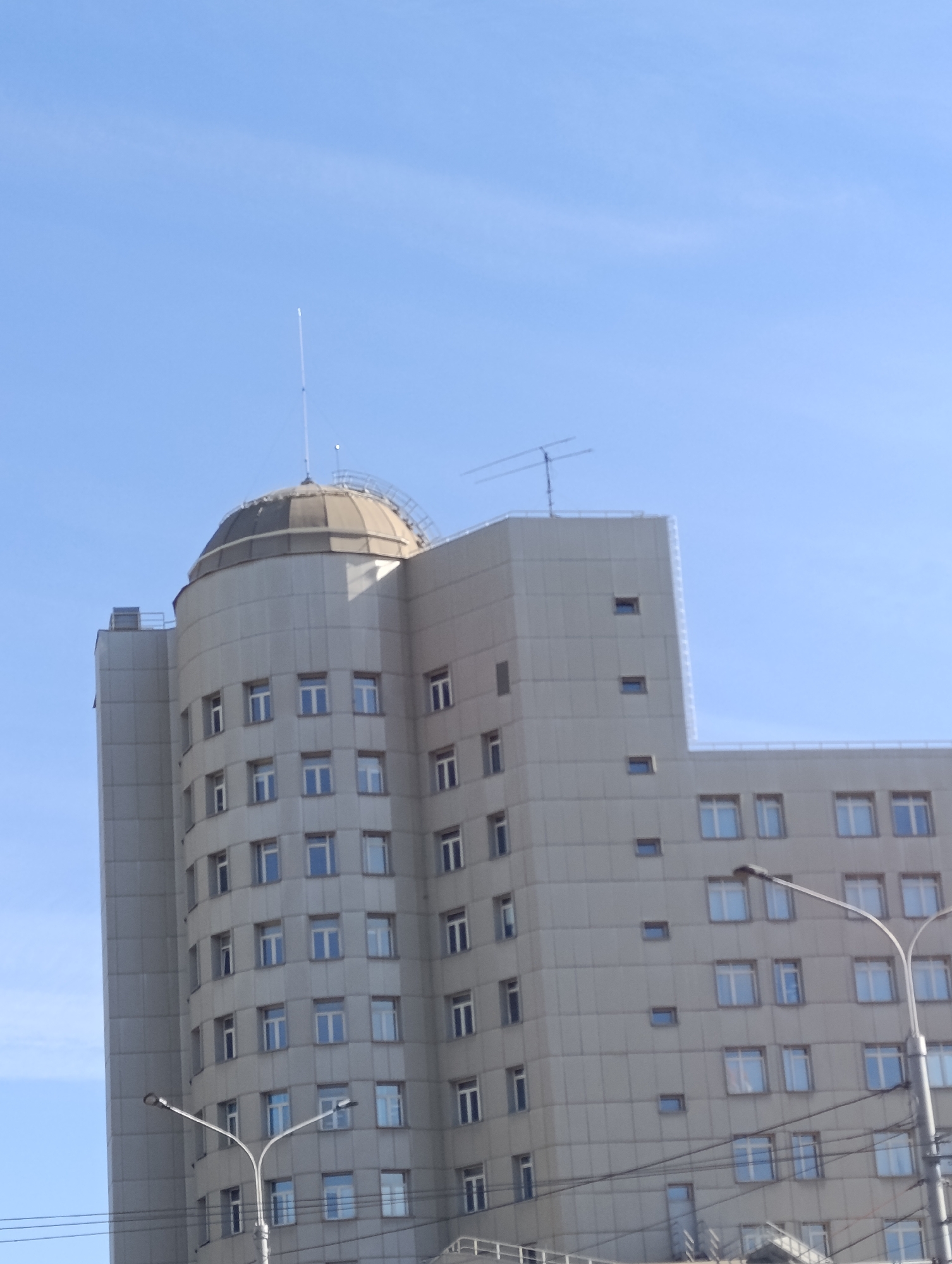 The image displays the Legislative Assembly of Krasnoyarsk Krai, located at Prospekt Mira 110, Krasnoyarsk, Russia, under a clear, bright blue sky. The multi-story building features a dominant light grey facade composed of large rectangular panels.
The left section of the building is cylindrical with a prominent domed roof, also grey. A slender, white antenna or lightning rod extends vertically from the dome's apex. A narrow perimeter walkway or railing is visible around the base of the dome. Windows on this curved section are rectangular, arranged in vertical columns, and appear to have blue-tinted or reflective glass.
To the right, the building extends with flatter, stepped sections. These sections also feature numerous rectangular windows, similarly tinted, arranged in regular rows and columns. Additionally, smaller, square-shaped windows are interspersed on some of the upper levels of the flatter sections. Another, wider Yagi-style antenna is mounted on a flat rooftop section to the right of the dome.
In the lower foreground, two light grey street light poles with bifurcated arms and double luminaires are partially visible, obscuring the building's lower levels. Multiple dark electrical or communication cables crisscross horizontally in the mid-ground, connected to the street light structures. No individuals or discernible actions are present in the scene. The lighting suggests daytime conditions with direct sunlight.