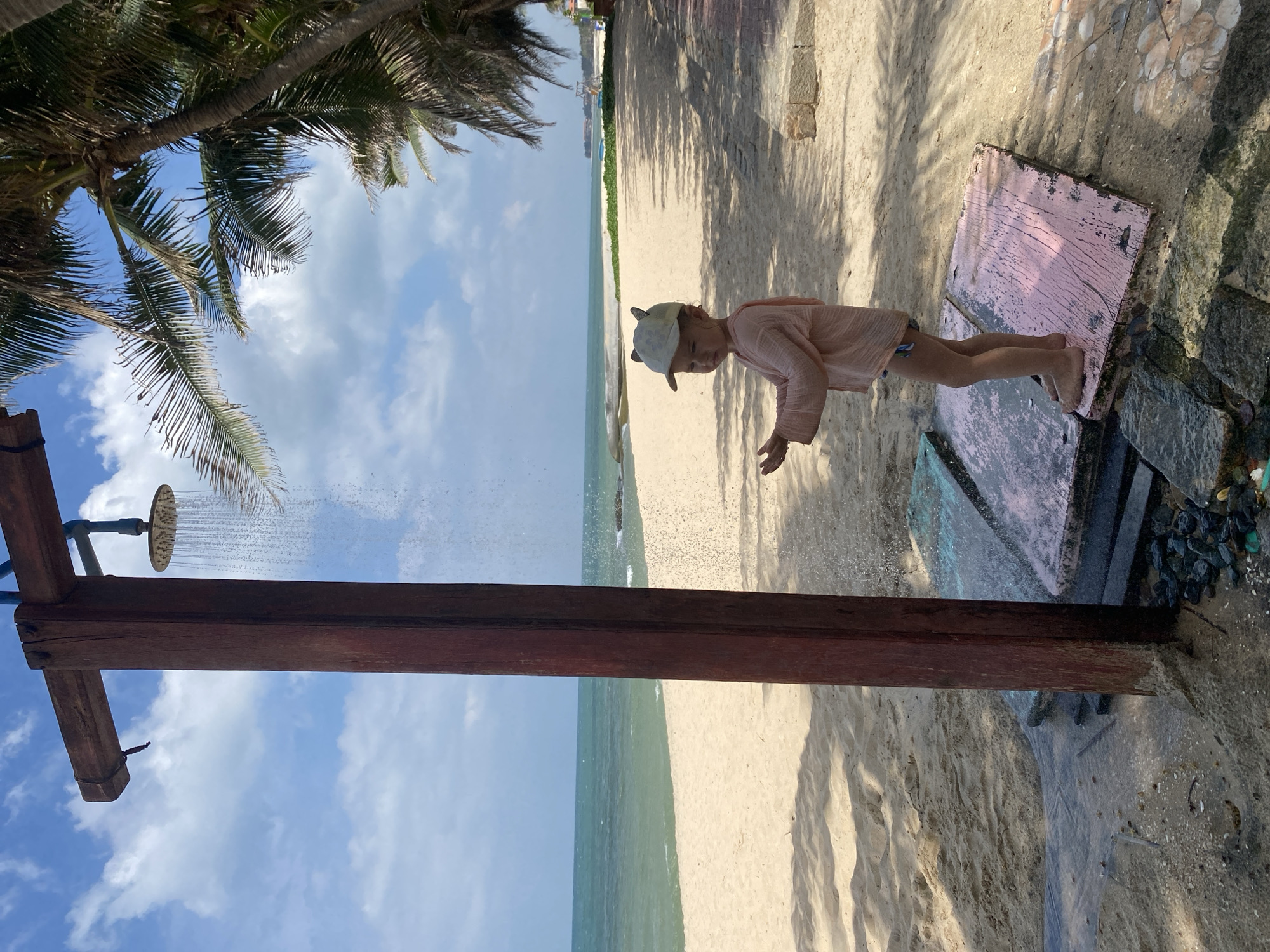 A young child, wearing a light pink long-sleeved shirt and a hat with cat ears, stands on a beach in Phu Thuy Ward, Vietnam, under an outdoor shower. The child's hands are outstretched as water cascades from the showerhead above. The beach has white sand and is bordered by palm trees on the right. The ocean is visible in the background, with calm, turquoise water meeting a sandy shoreline. The shower structure is made of wooden posts. The child is barefoot and standing on colorful, weathered wooden platforms, which are placed on the sand. The ground around the shower appears to be a mix of sand, stones, and some water runoff. The sky is blue with scattered white clouds.