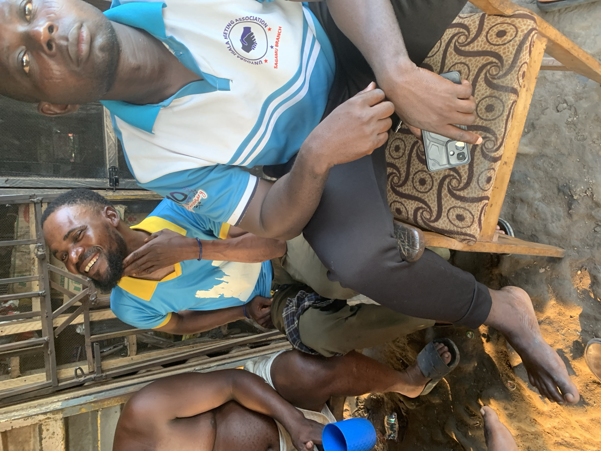 This is a medium shot, eye-level outdoor photograph of a group of men sitting on chairs and on the ground. The image is taken in Shagamu, Nigeria. The setting appears to be a casual gathering, possibly a marketplace or a community space, with a rustic feel.
In the foreground, a man is seated on a wooden chair with a patterned cushion. He is wearing a white and blue polo shirt with a logo that reads "UNYOGBA IGALA MEETING ASSOCIATION SAGAMU BRANCH." He is holding a dark-colored smartphone in his right hand, with his finger resting on the screen. His legs are crossed, and he is wearing dark pants.
To the left of the seated man, another man is sitting on the ground. He is wearing a bright blue t-shirt with a yellow collar, and khaki pants. He has a beard and is laughing heartily, with his hand touching his neck. Next to him, a blue plastic cup is placed on the ground.
Further to the left, another man is partially visible, seated on the ground. He is bare-chested and wearing light-colored shorts.
In the background, there is a metal gate or fence, suggesting the area is either enclosed or adjacent to a structure. The ground is made of dirt and sand, with some debris scattered around. The overall lighting suggests it is daytime.