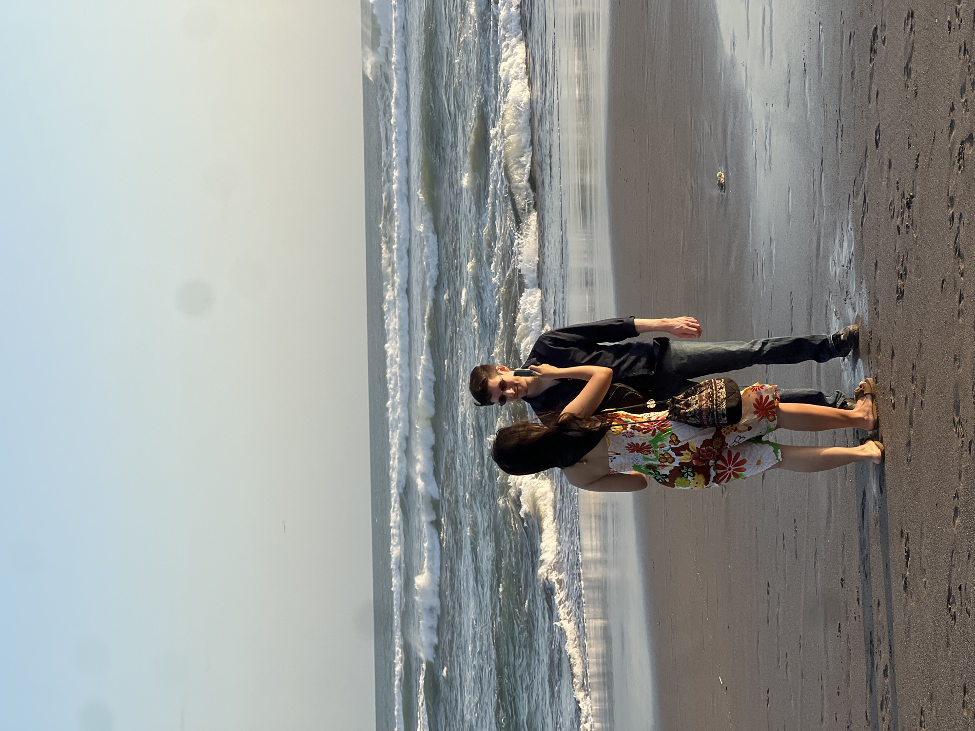 An over-the-shoulder shot captures a young couple on a sandy beach with ocean waves in the background. The woman, facing away from the camera, wears a floral sundress and holds up a smartphone, seemingly taking a picture of the man. She has dark hair and wears sandals. The man, facing her, wears a dark button-down shirt, jeans, sunglasses, and sneakers. He smiles at the woman as she takes his picture. The beach sand shows footprints, and a small object, possibly a shell, sits in the wet sand near the man's feet. The ocean exhibits multiple lines of breaking waves, and the sky above is a pale blue. The scene appears to be in North Kuta, Indonesia, based on the context provided.