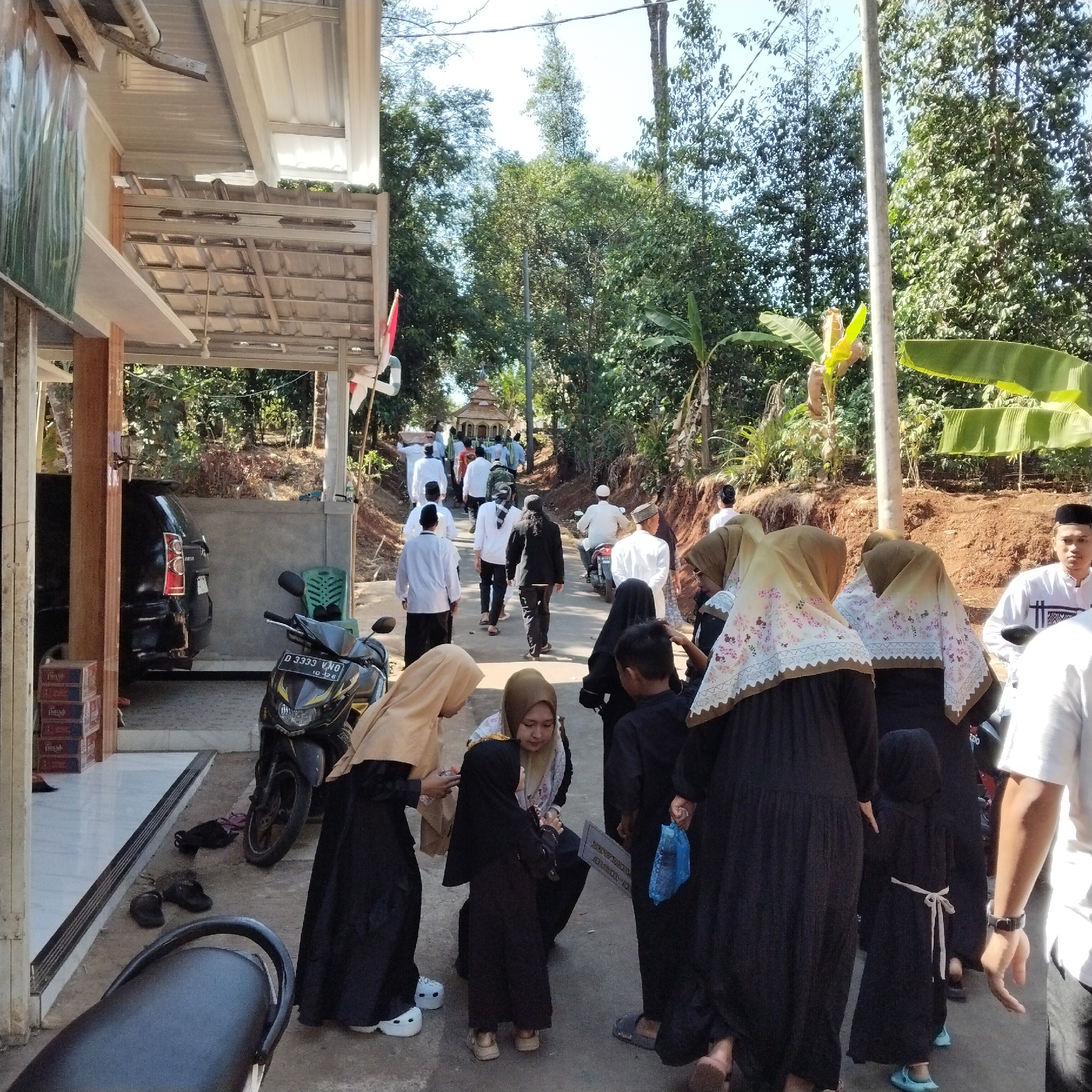 The image captures an outdoor scene in Pamulihan, Indonesia, with a focus on a group of people, many wearing traditional Islamic attire. In the foreground, several individuals are gathered, including women in hijabs and long dresses. To the left, a black SUV is parked under a covered structure. A motorcycle is also present. The background shows a dirt road leading uphill, flanked by trees and vegetation, suggesting a semi-rural or suburban environment. A small, ornate building is visible further up the road. Several people are walking up the road, some in white clothing and skullcaps, possibly attending a religious gathering or procession. The overall atmosphere appears to be one of community and religious observance.
