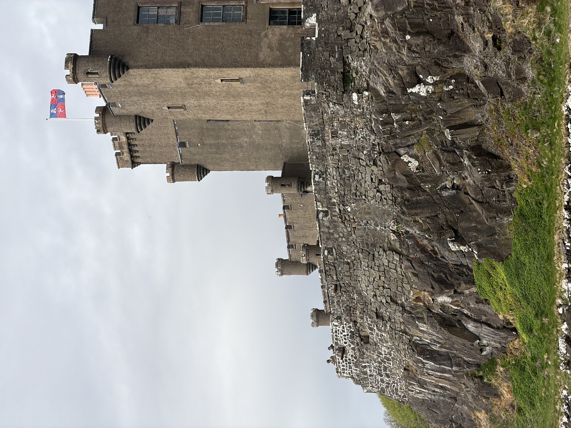 The image depicts Dunvegan Castle, located in Dunvegan, United Kingdom. The castle is built on a rocky promontory overlooking a body of water, likely the sea. The castle itself is constructed of stone and features several towers and turrets. A flag is visible atop one of the towers. The surrounding environment consists of rocky cliffs and sparse vegetation. The sky is overcast. There is a hint of green in the bottom right corner, possibly vegetation. There are two figures standing on the outer castle wall.