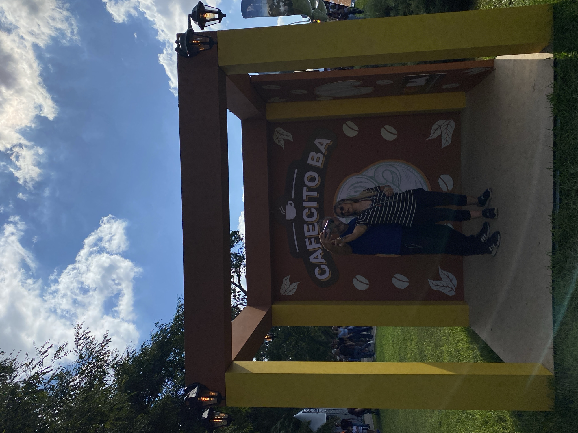 The image shows a "Cafecito BA" photo booth in Buenos Aires, Argentina. It is a rectangular structure with brown and yellow beams. The back panel is brown with white coffee bean and leaf designs, and the words "CAFECITO BA" in white lettering. Two people are posing for a photo within the booth. A woman is wearing a blue shirt and black pants and holding a phone. A man is wearing a black and white striped shirt and black pants. The background shows a grassy area and other people, suggesting the structure is in a park or public space. The sky is blue with scattered clouds. Street lamps are attached to the brown sections of the structure.