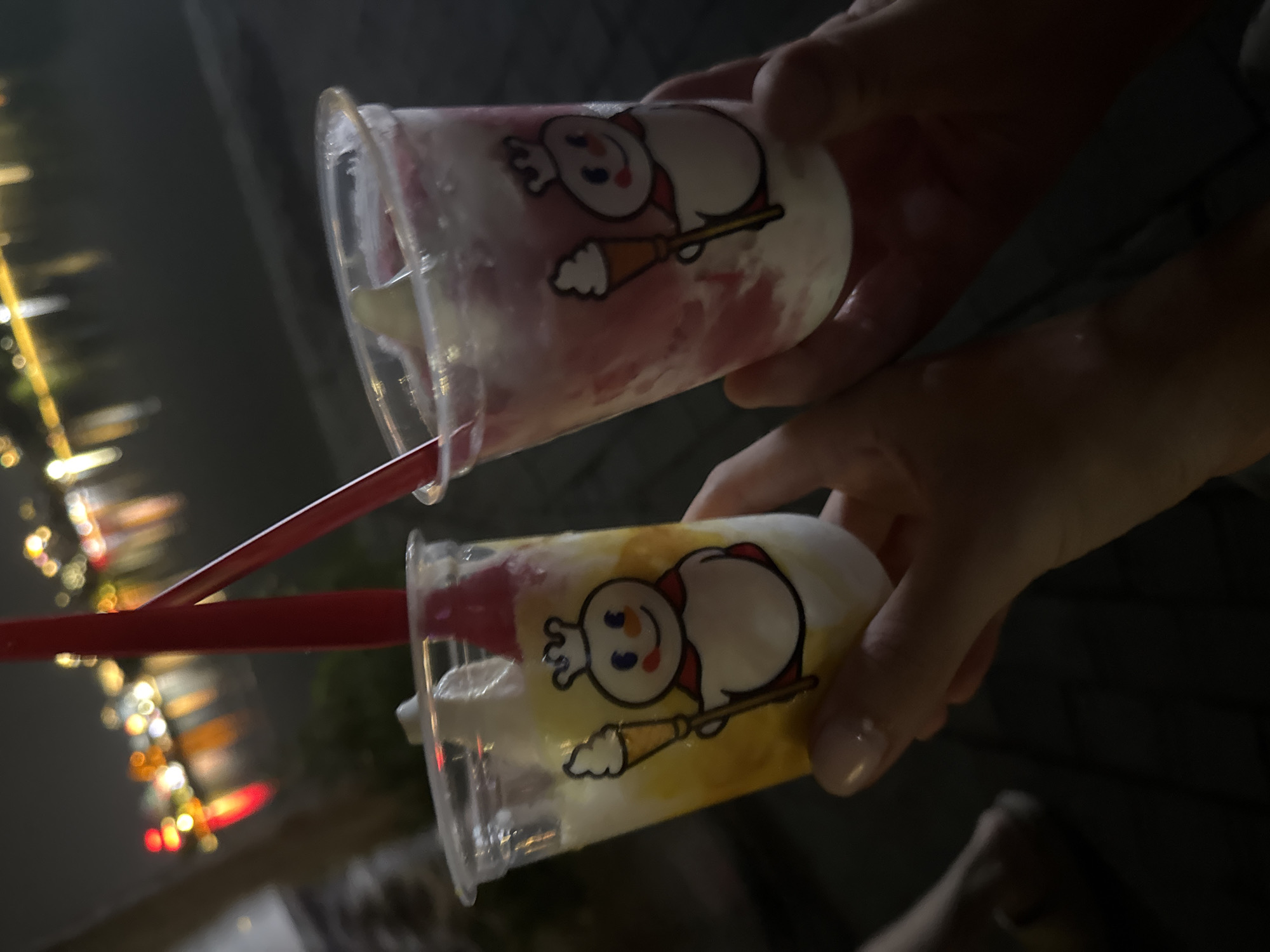 In a nighttime outdoor setting, two hands hold two cups, each containing a blended beverage of different colors. The cup closer to the bottom of the frame contains a yellow-tinted beverage, while the upper cup has a pinkish-red hue. Both cups are adorned with a cartoonish snowman logo. Red straws extend from the drinks. The background displays blurred lights, possibly of a city skyline. The image was taken in Hà Nội, Vietnam.