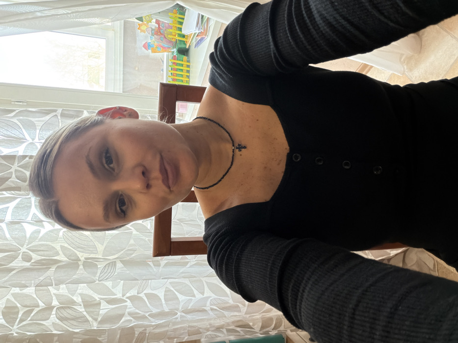 A light-skinned adult female is depicted in a selfie, looking directly at the camera with a slightly pursed expression. Her light-colored hair is pulled back. She wears a black long-sleeved top with a square neckline, featuring three visible buttons down the front, and a black beaded necklace with a dark cross pendant. A small stud earring is visible in her right ear. She appears to be seated, with a wooden chair backrest visible behind her. The background indicates an indoor setting in the city of Chaikovsky, Russia. Light-colored, sheer curtains with a repeating leaf pattern flank a window. Outside the window, bare tree branches are visible under natural light. On a surface below the window, colorful objects resembling children's crafts or toys, including a multi-colored castle-like structure and a yellow fence, are arranged alongside white papers.