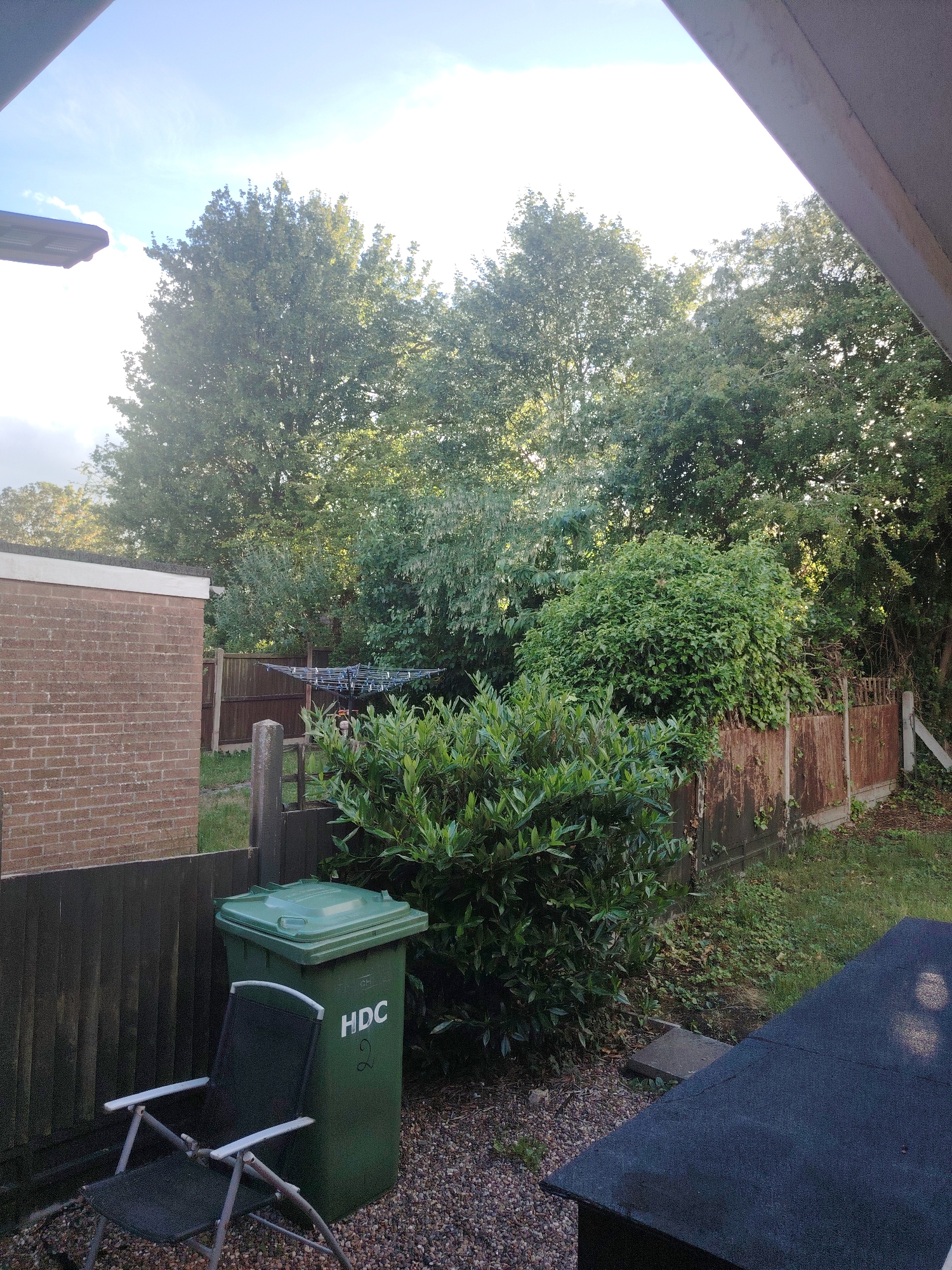 This outdoor image depicts a residential backyard or garden scene, likely in Huntingdonshire, United Kingdom, given the provided context and the "HDC" marking on the bin.
The scene is dominated by lush green foliage, with tall trees forming a dense canopy across the background and mid-ground. Bright sunlight filters through the leaves and illuminates parts of the scene, indicating it is daytime, possibly late morning or afternoon. The sky is clear with a few wispy clouds.
In the foreground, a dark green wheelie bin stands prominently, bearing the white text "HDC" and the number "2". Leaning against the bin is a black and silver folding garden chair. The ground in this area is covered in small gravel. A dark wooden fence runs from the left, separating the gravel area from a patch of grass.
On the left side of the frame, the red-brick wall of a building with a flat roof is visible. Further back in the mid-ground, behind thick green bushes, a rotary clothesline is partially visible. To the right, a weathered and rusty metal fence, partially obscured by overgrown vegetation, marks a boundary. A concrete fence post is also present.
No people are visible in the image. The setting conveys a quiet, typical domestic outdoor space.