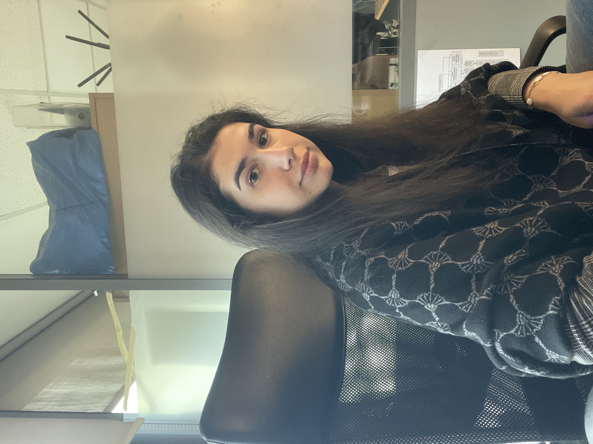 A young woman sits in a black office chair, with long dark hair and fair skin. She wears a dark patterned jacket over a collared shirt, and a bracelet on her wrist. The setting appears to be an office environment. A white wall behind her features a shelf with a blue object, and a metal cabinet. A piece of paper with a diagram is visible behind the woman. The location in the city of Sofia, Bulgaria cannot be determined.