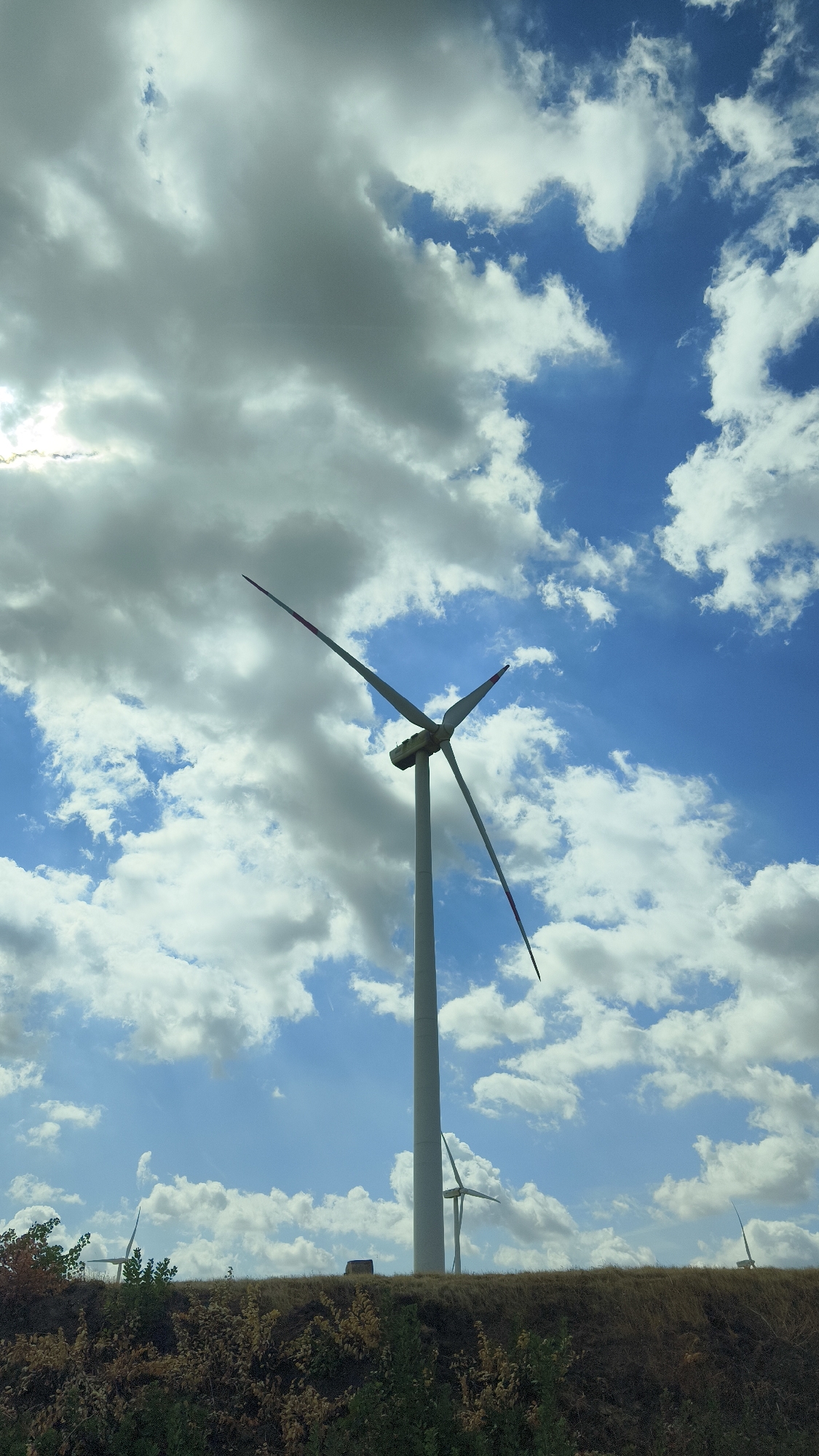 A full shot captures a wind turbine against a backdrop of a dynamic sky. The turbine stands tall, its three blades positioned at an angle, one slightly ahead of the others. The sky is a vibrant blue, dotted with fluffy white clouds and some darker, more diffused clouds, suggesting an overcast or partly cloudy day. The base of the turbine is not visible, as the shot is framed from a lower vantage point, looking up. In the distance, a few more wind turbines are visible, smaller in scale, suggesting a wind farm. The foreground shows a grassy embankment covered in dry grass and some green foliage, with a few scattered trees on the left. A small dark structure, possibly a shed or building, is barely visible on the embankment between the main turbine and the distant ones. The image appears to be taken from a moving vehicle, as evidenced by a faint horizontal line across the top of the frame, possibly a reflection or artifact from a window. The scene depicts a rural landscape with renewable energy infrastructure in Kisigmánd, Hungary.