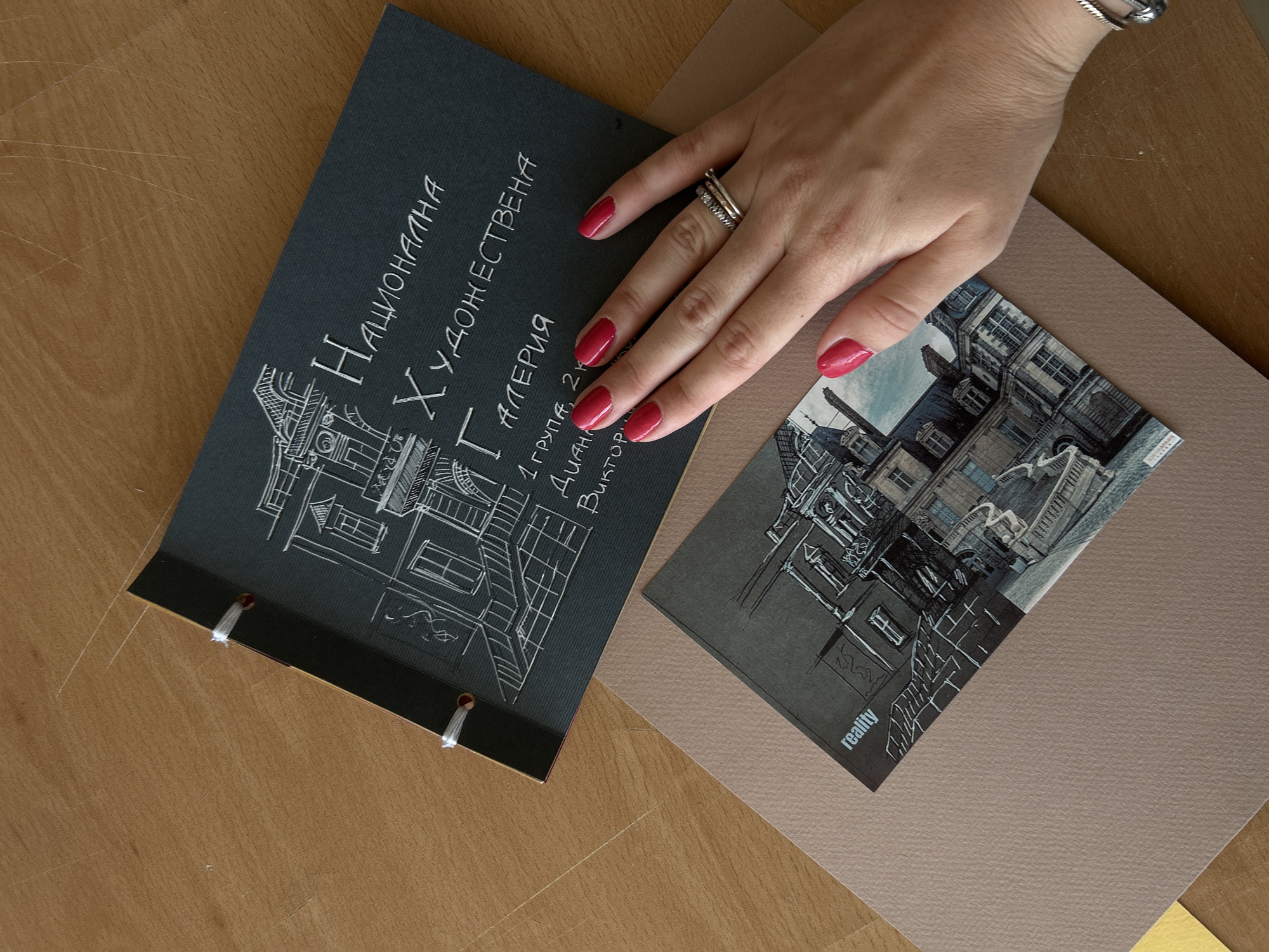 A hand with bright pink nail polish rests on a dark grey notebook, which is lying on a wooden surface. The notebook has white text and drawings of a building. Below the notebook is a smaller, printed image of a building and a tan piece of paper. The notebook is bound with white string. The image does not contain any information about the city of Sofia, Bulgaria.