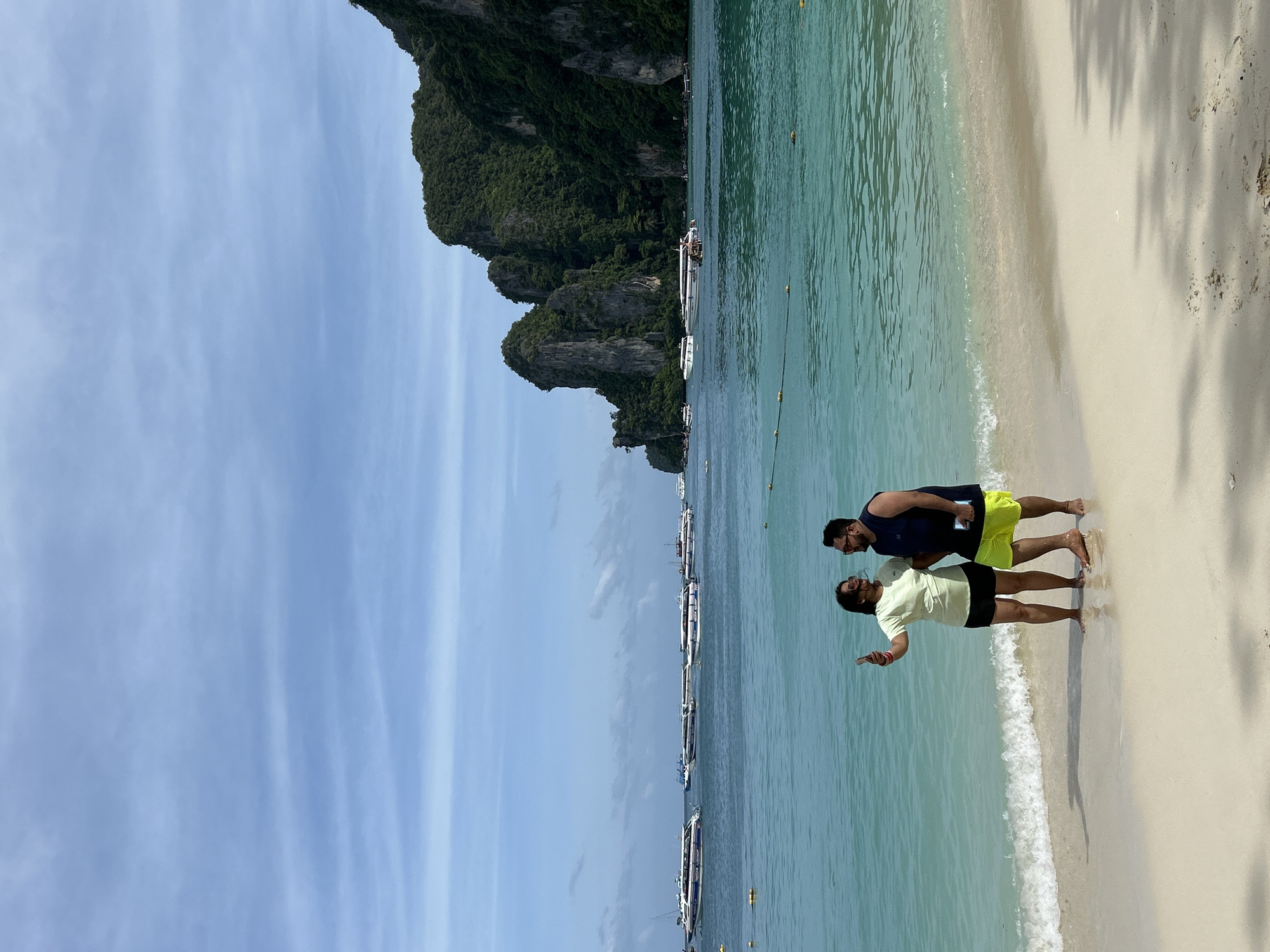 An overhead, medium shot captures a couple taking a selfie on a sandy beach in Ao Nang, Thailand. The woman, wearing a light green t-shirt and black shorts, holds a smartphone to her left as she looks at the camera. Beside her, a man in a navy blue sleeveless shirt and bright yellow shorts looks at her. The pair stands at the edge of the turquoise water, with small waves lapping at their feet. In the background, several long-tail boats are anchored in the calm sea, with a backdrop of lush green, rocky cliffs. The sky above is a pale blue with scattered white clouds.