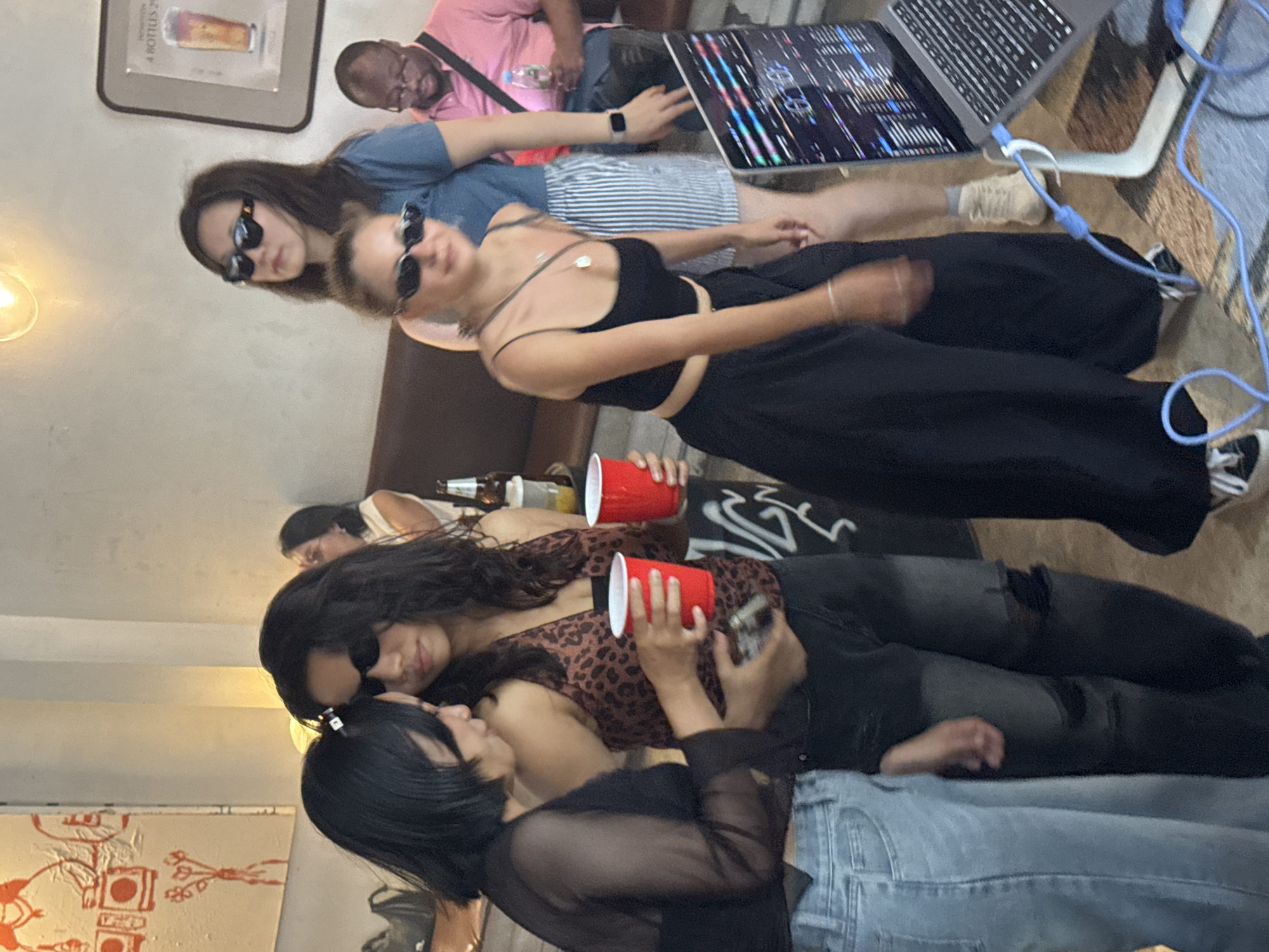 The image depicts a group of people socializing in what appears to be an indoor venue in Chiang Mai City Municipality, Thailand. In the foreground, two women are standing close together. The woman on the left has short black hair and is wearing a sheer black top and light blue jeans. She is holding a phone. The woman next to her has long dark hair, sunglasses, and is wearing a leopard-print top and dark jeans. She is holding two red plastic cups. Behind them, a woman with blonde hair and sunglasses is wearing a black tank top and black pants, and is holding a drink. Further in the background, a woman in a blue t-shirt and striped shorts is also present, along with a man in a pink polo shirt. A laptop is visible in the lower right corner, displaying a graphical interface. The environment suggests a casual gathering, possibly a bar or event space, with artwork visible on the wall and dim lighting.