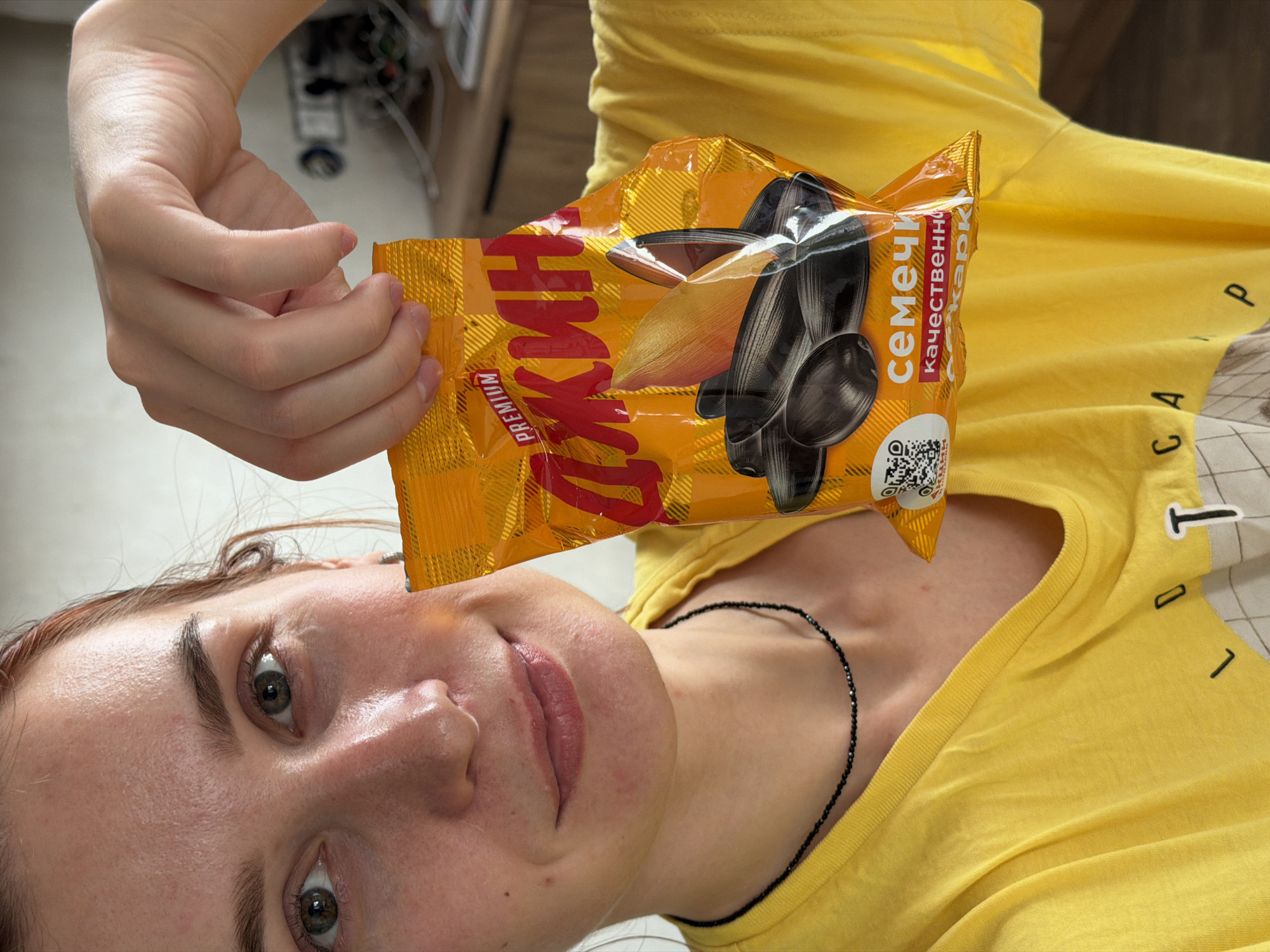 A person is holding a yellow package of sunflower seeds close to their face. The person is wearing a yellow t-shirt and a black beaded necklace. The package of sunflower seeds is labeled with Russian text and a QR code. The person is in an indoor environment. It is not possible to confirm the location as Chaikovsky, Russia based on the image alone.