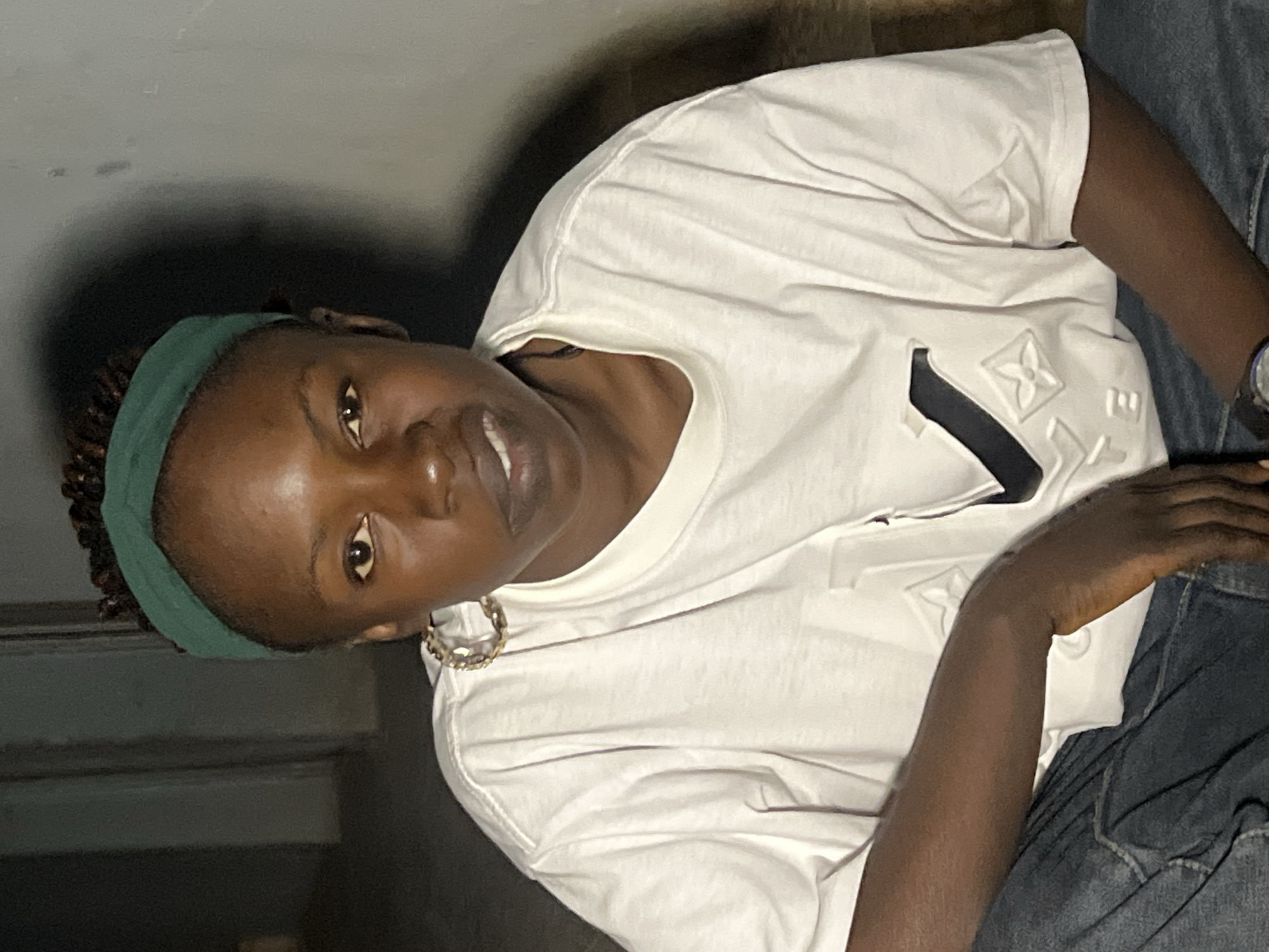 A medium shot captures a young Black woman wearing a white t-shirt and dark denim jeans, seated indoors. She has dark skin, her hair styled in braids and adorned with a green headband. A large hoop earring is visible on her left ear. Her facial expression is slightly asymmetrical, with her mouth open and teeth showing. The background appears to be a dimly lit interior with textured walls. The woman's left arm rests on her lap, and her right arm is bent. The setting suggests an informal, domestic environment within the city of Jos, Nigeria.